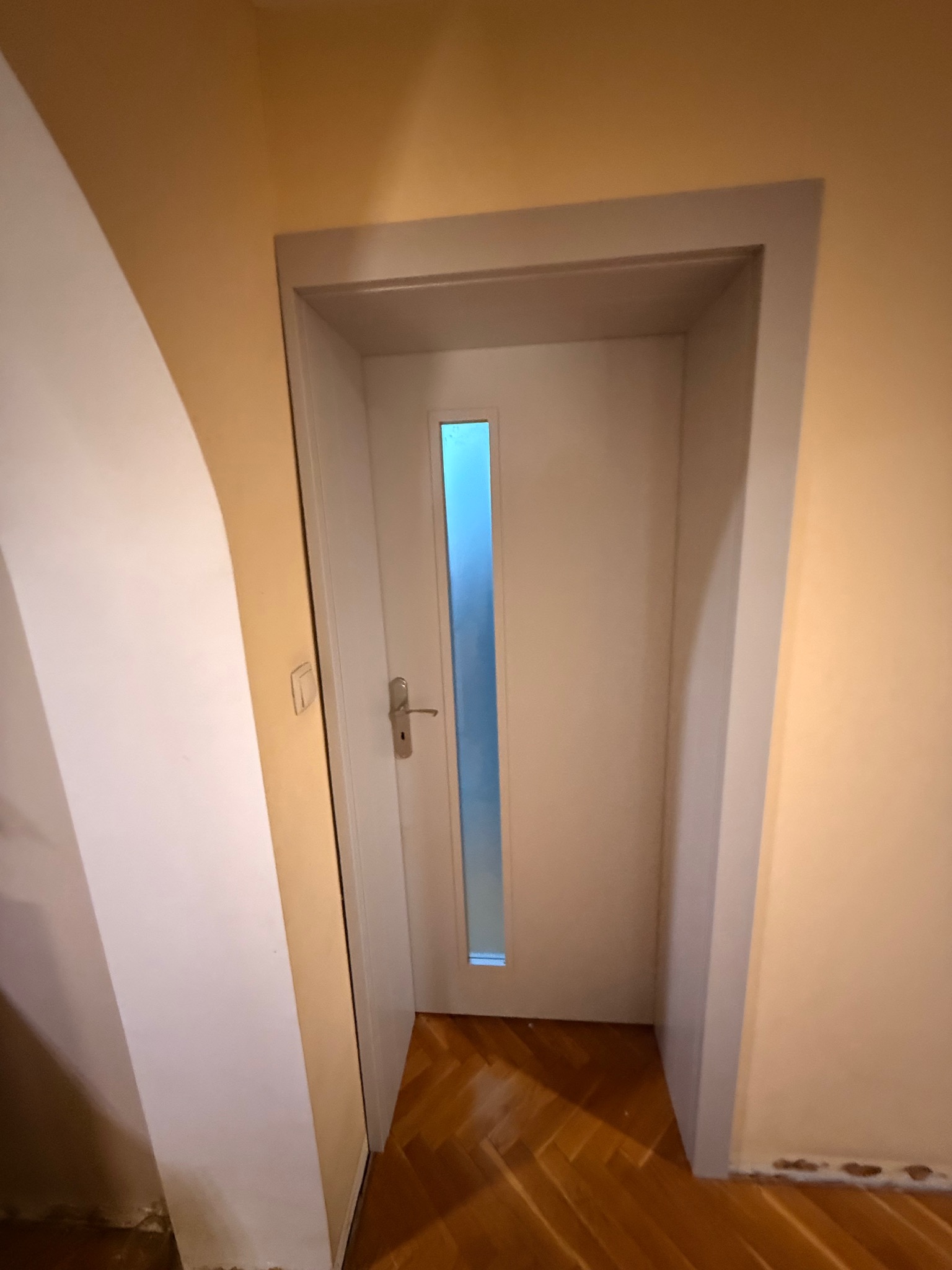 Białe drzwi wewnętrzne z pionowym przeszkleniem, osadzone w szarej ościeżnicy, na tle beżowej ściany i parkietu. Widoczny fragment białej ściany po lewej stronie.