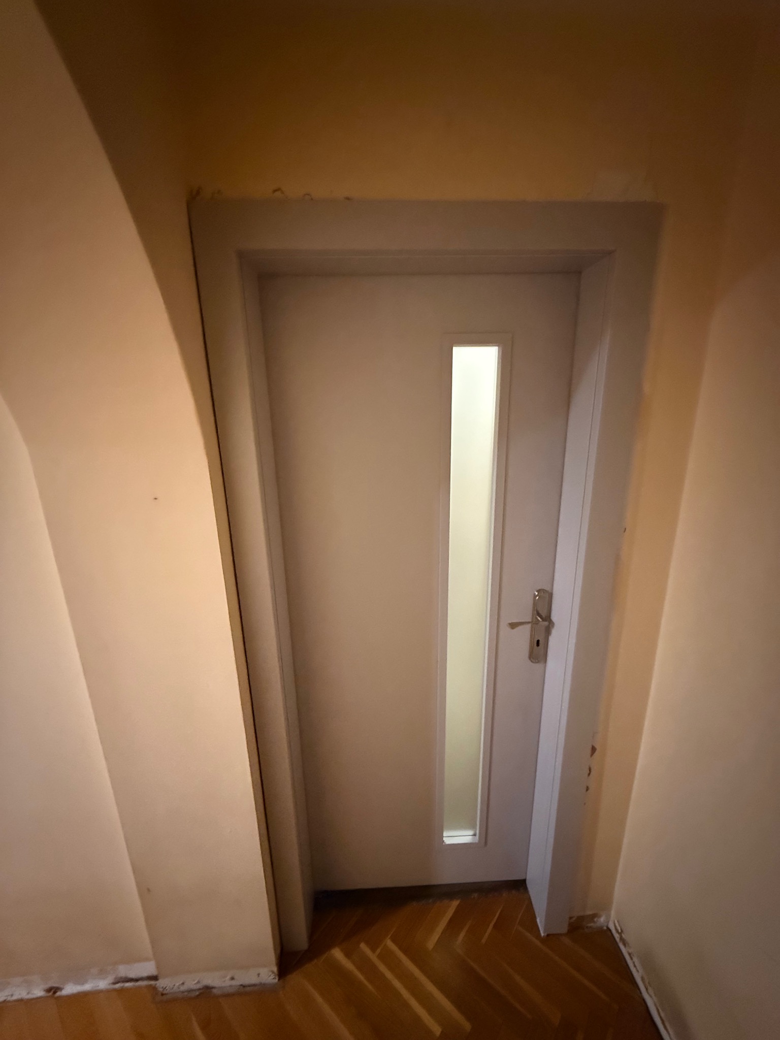 Białe drzwi wewnętrzne z przeszkleniem, zamontowane w ościeżnicy. Widoczna podłoga z parkietu i fragment ściany w odcieniach beżu. Ujęcie z bliska, lekko z dołu.
