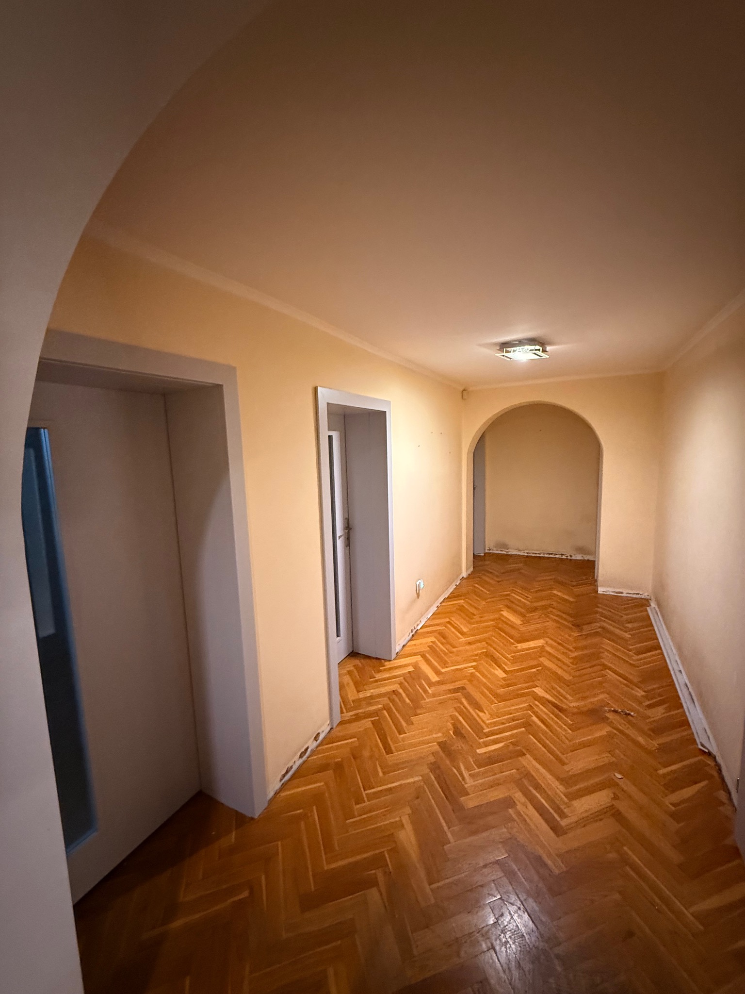 Korytarz z parkietem ułożonym w jodełkę, widoczne dwie pary drzwi i łukowate przejście. Ściany w odcieniach beżu, sufit biały z lampą. Widok z perspektywy wejścia.