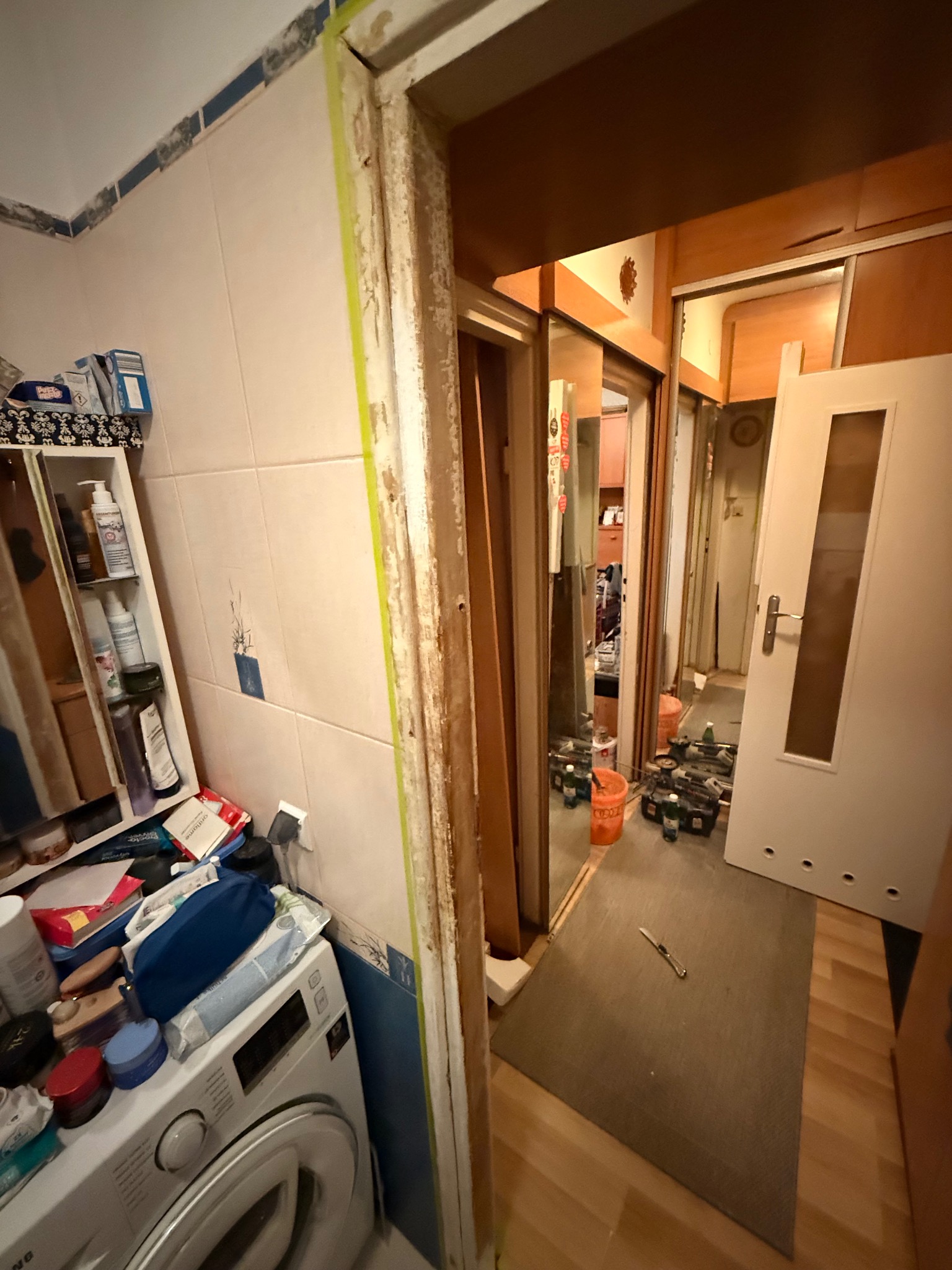 Przygotowanie do remontu łazienki: usunięte futryny, zabezpieczone krawędzie taśmą malarską, widoczny bałagan i narzędzia. W tle widoczny korytarz z drzwiami.