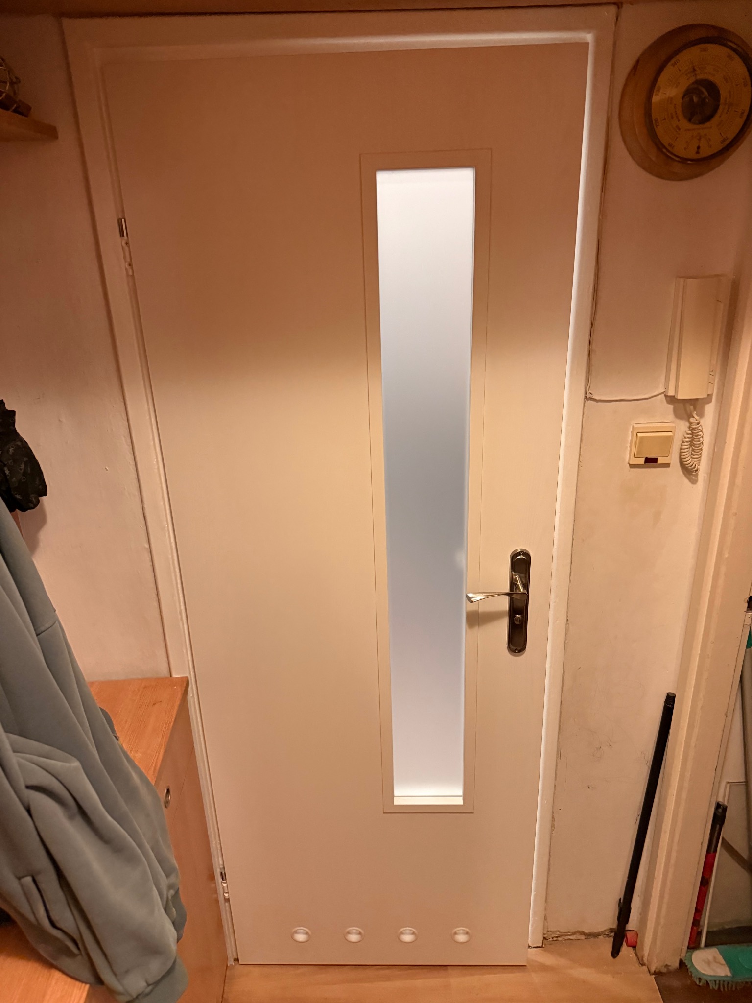 Białe drzwi wewnętrzne z przeszkleniem w pionowym pasie, zamontowane w mieszkaniu w Warszawie. Widoczny fragment ściany z barometrem i włącznikiem światła.