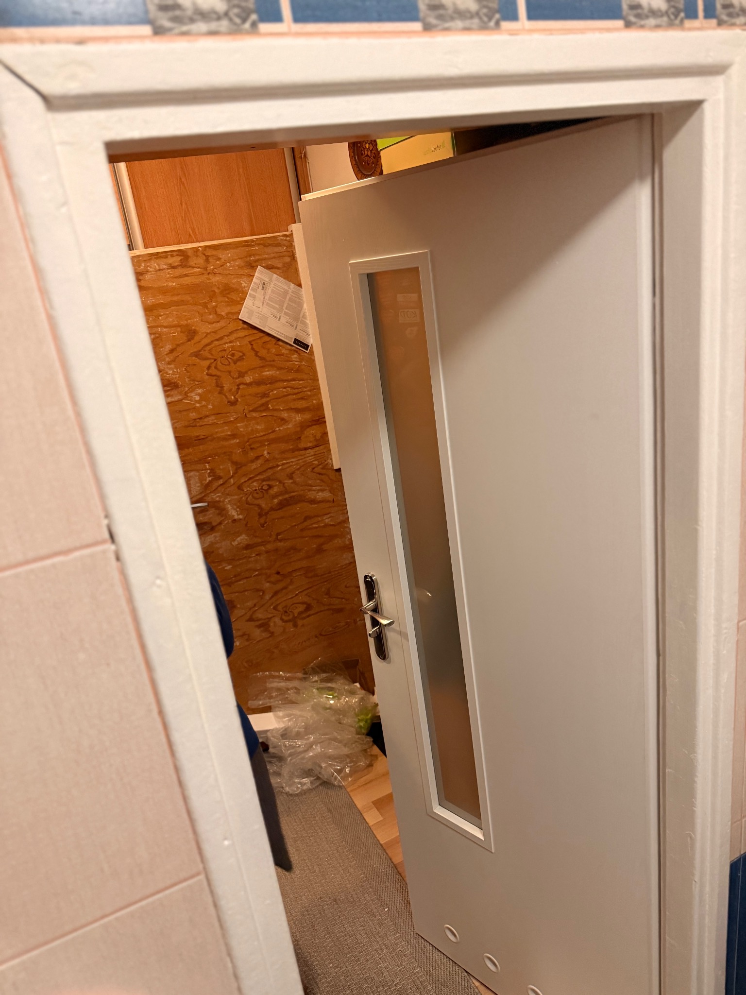 Białe, częściowo otwarte drzwi z szybą, widoczne z perspektywy wnętrza. W tle fragment ściany z boazerią i podłoga. Ujęcie z bliska, z lekkim pochyleniem.