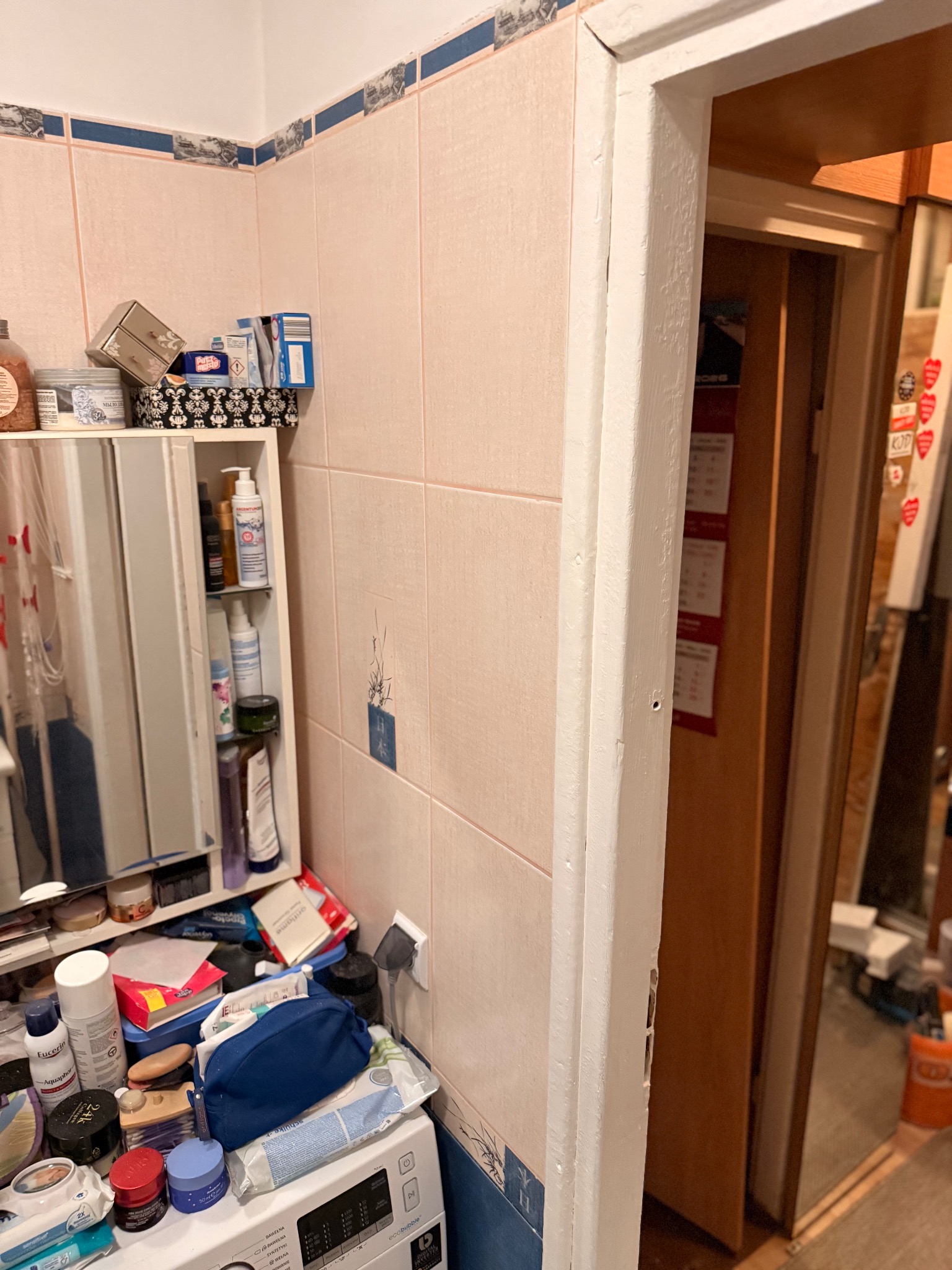 Łazienka przed remontem: różowe płytki z dekoracyjnym paskiem, biała ościeżnica drzwi z ubytkami farby, pralka z kosmetykami na wierzchu i szafka z lustrem.