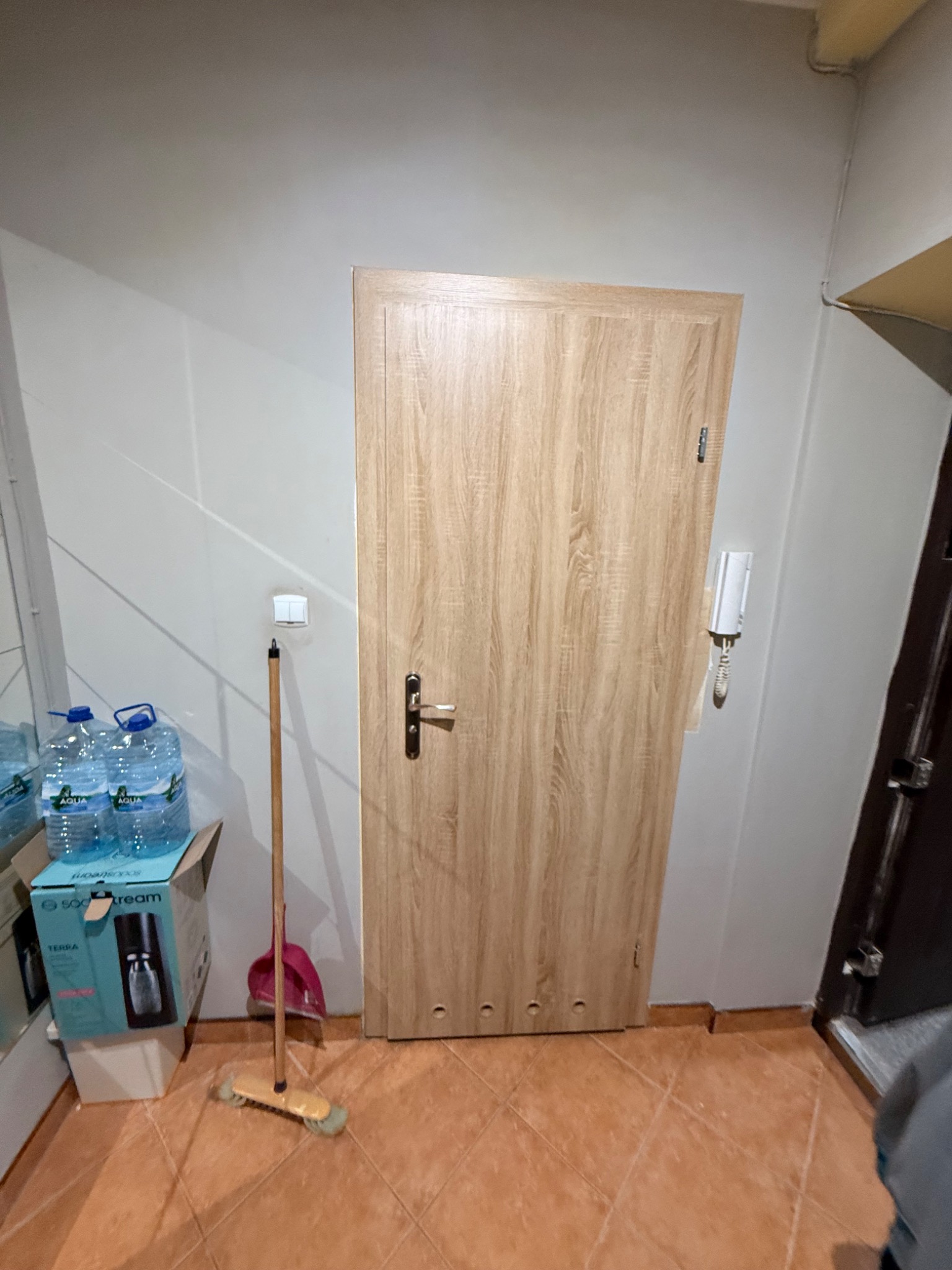 Jasna, drewnopodobna okleina drzwi wewnętrznych w korytarzu. Widoczny domofon na ścianie, brązowa podłoga i fragment lustra po lewej stronie.
