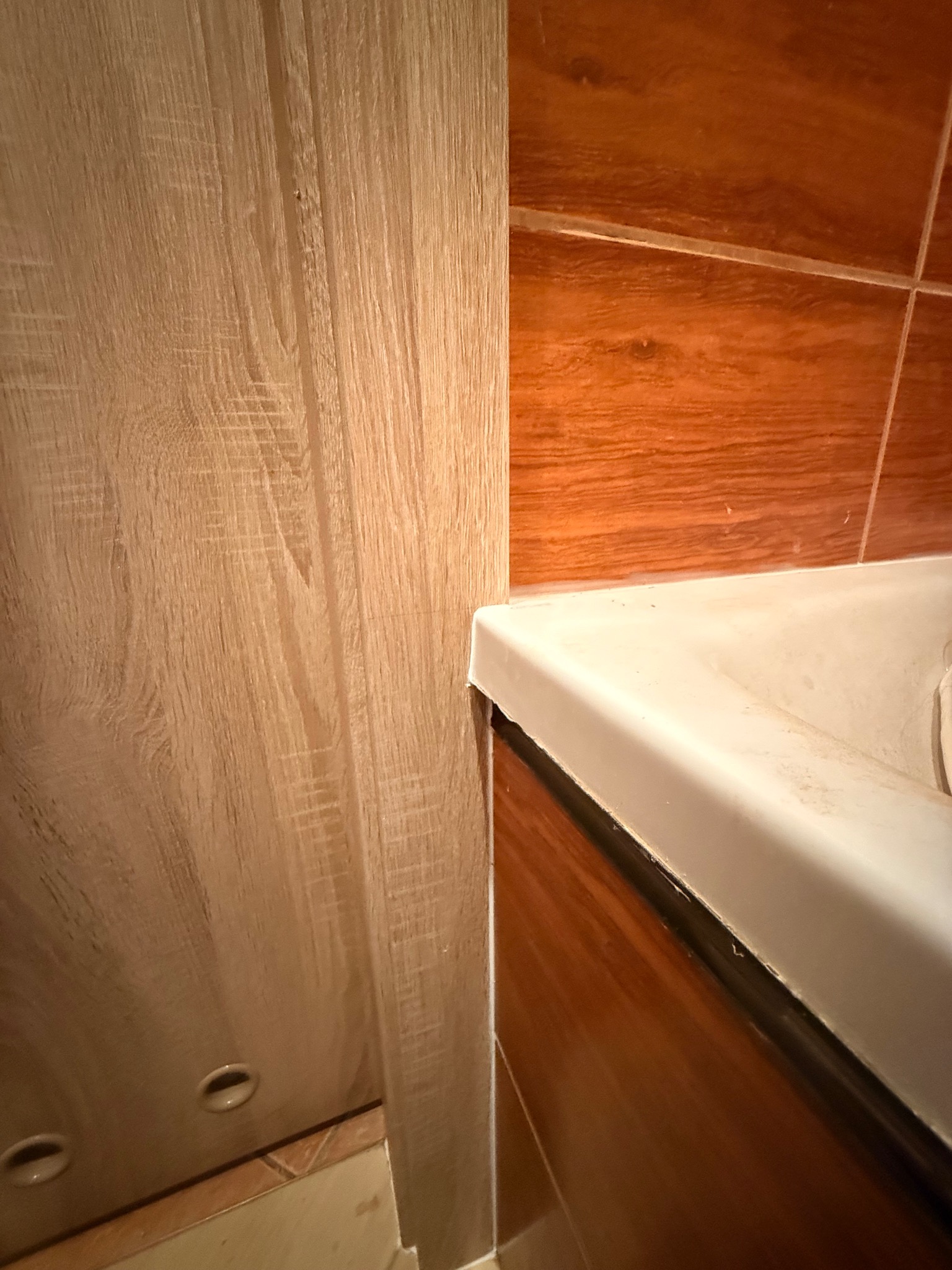 Fragment wykończonej łazienki: brązowe płytki, umywalka z drewnopodobną szafką i drewnopodobne wykończenie ściany z widocznymi otworami na akcesoria.