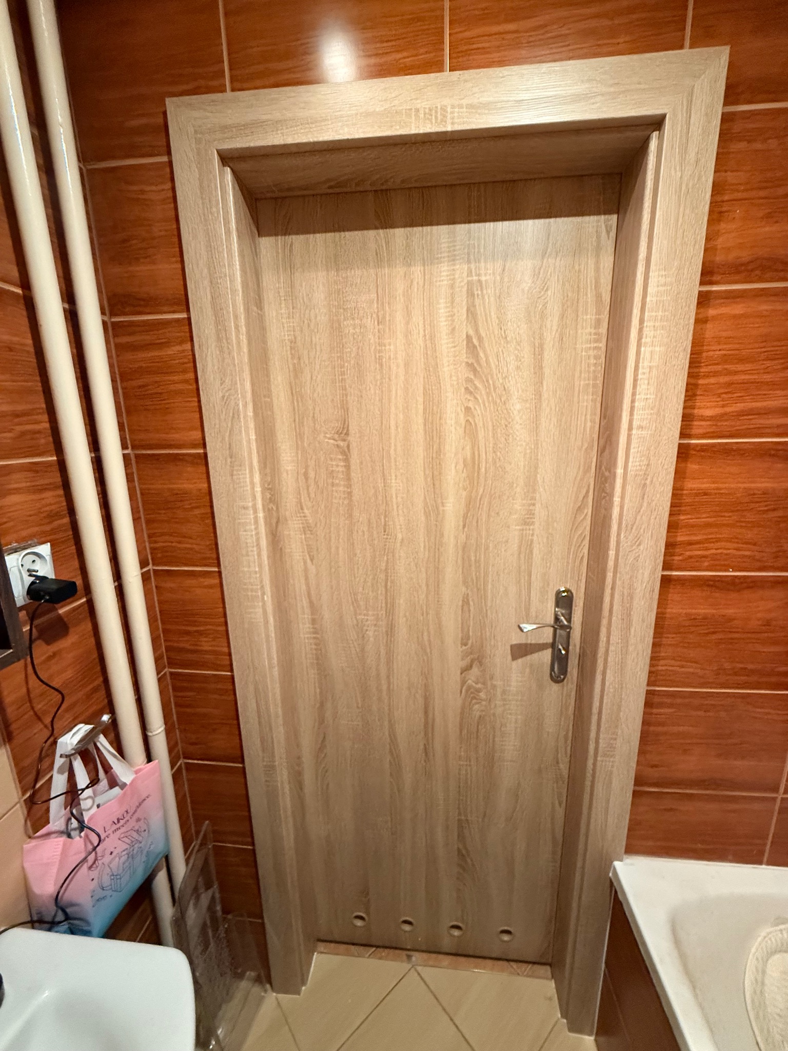 Jasna, drewnopodobna okleina drzwi wewnętrznych w łazience z brązowymi płytkami. Widoczne rury i fragment umywalki. Drzwi z klamką, zamontowane w ościeżnicy.