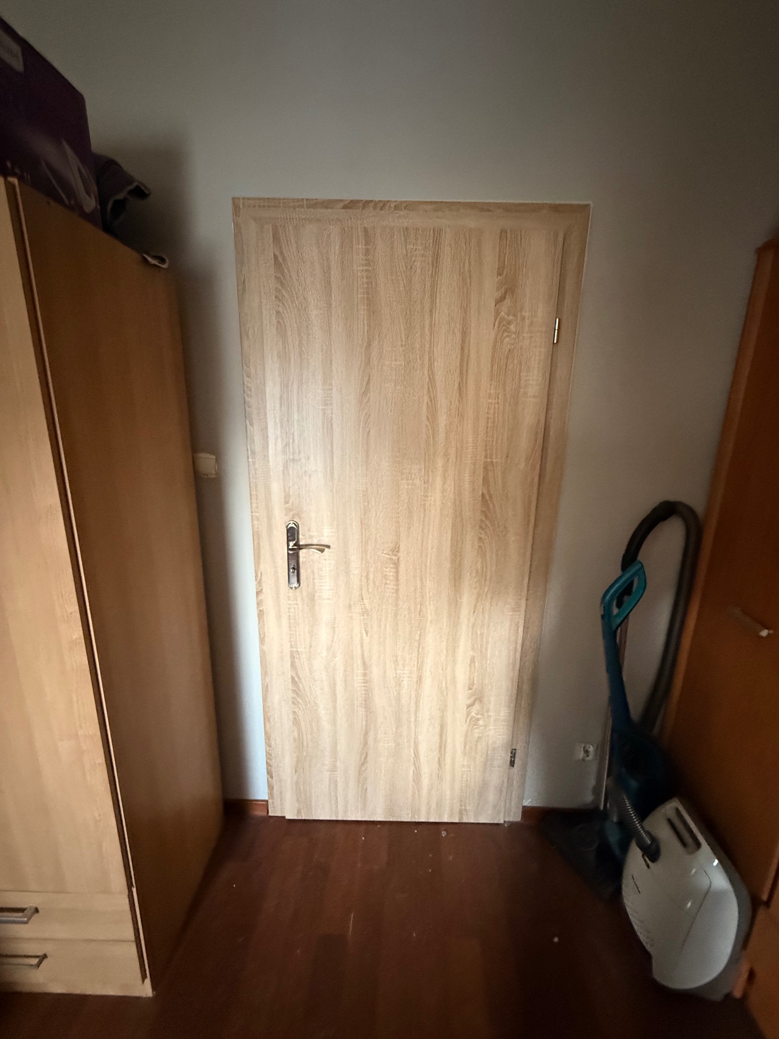 Jasne, drewnopodobne drzwi wewnętrzne z klamką, zamontowane w pokoju. Obok widoczny odkurzacz i fragmenty szaf. Ujęcie z poziomu podłogi.