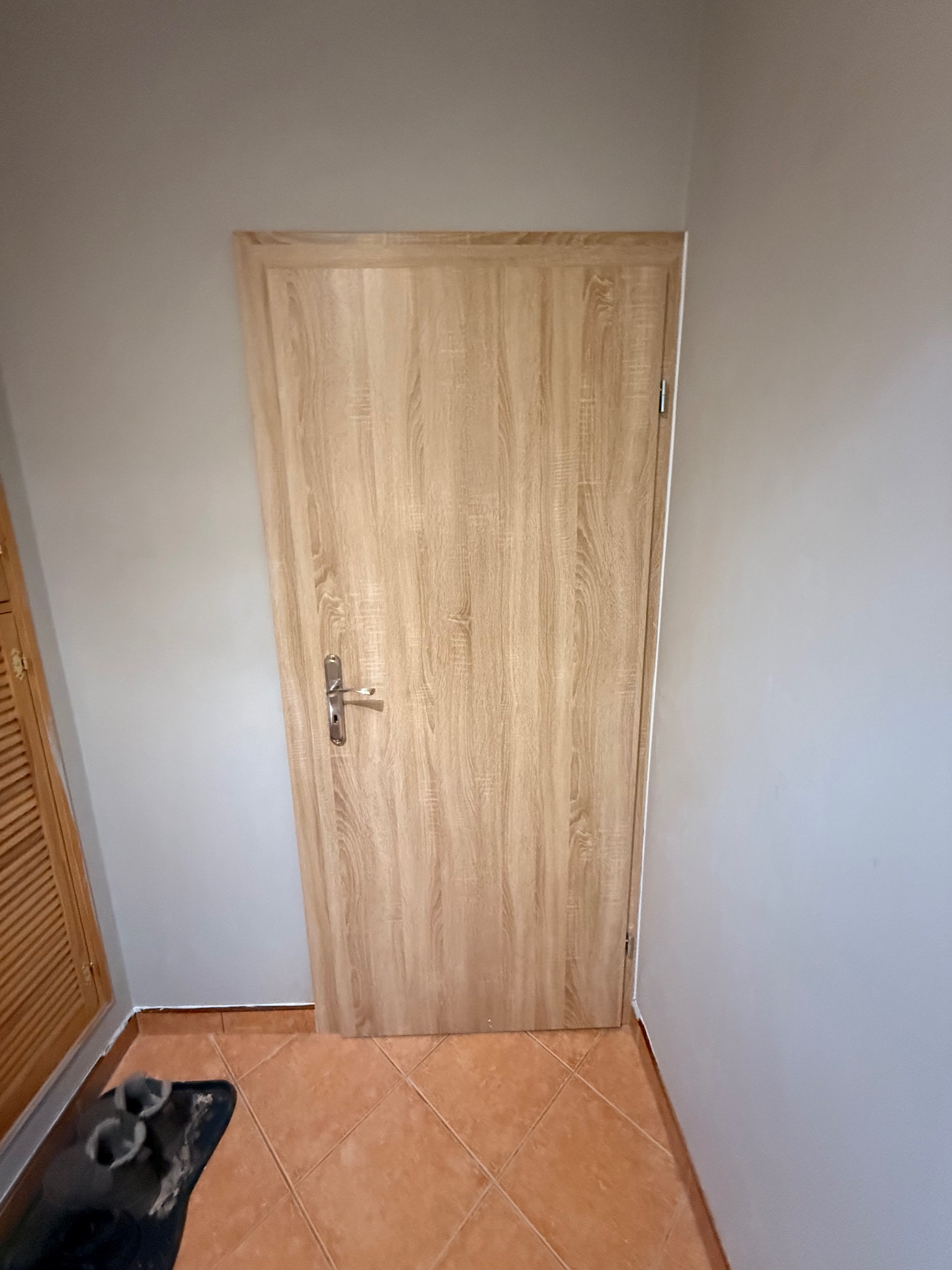 Zamknięte, drewnopodobne drzwi wewnętrzne z srebrną klamką, na tle szarej ściany i pomarańczowej podłogi z płytek. Widoczny fragment szafy z żaluzjowymi drzwiami.