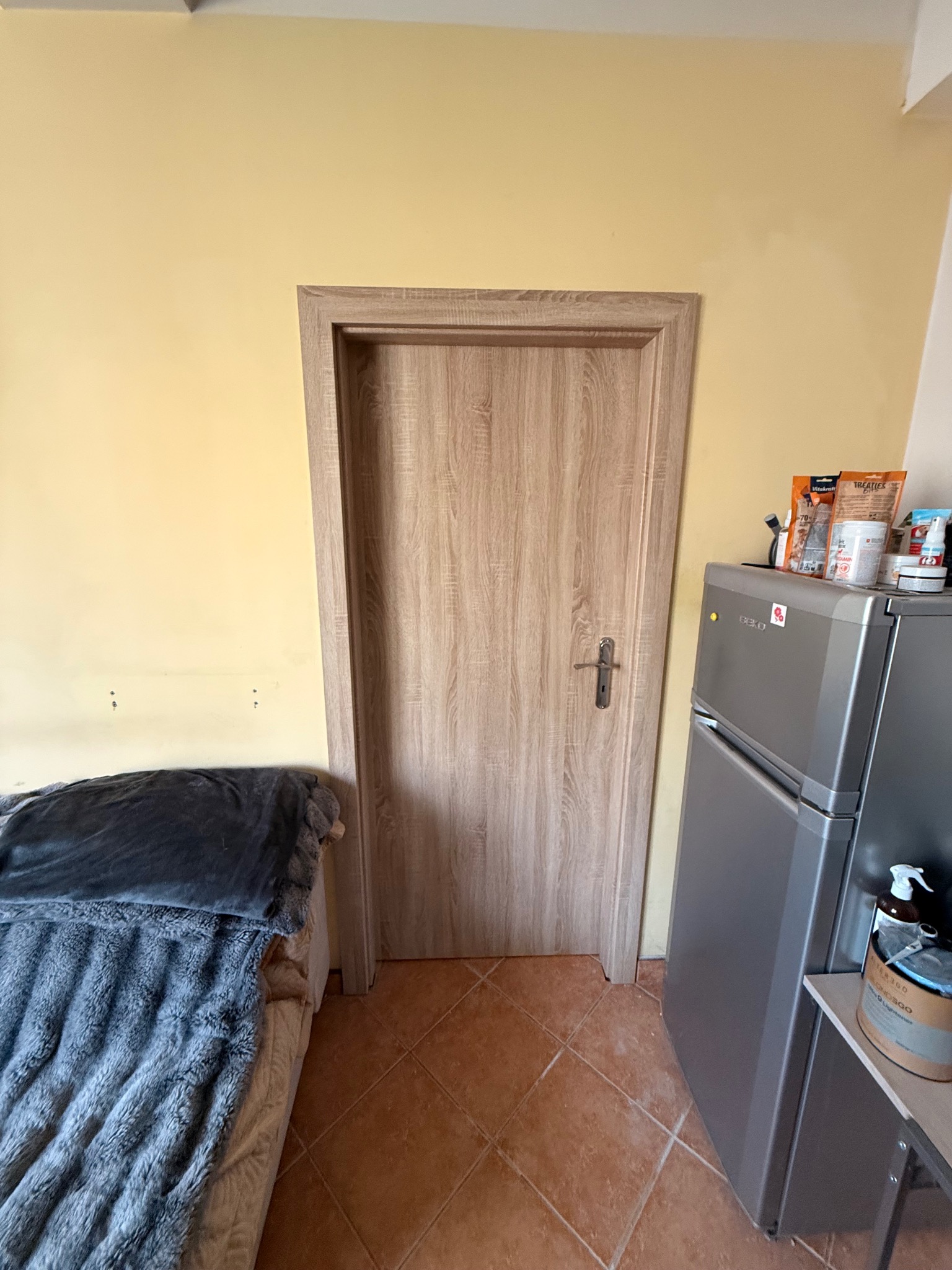 Zamknięte, drewnopodobne drzwi wewnętrzne z klamką, na tle żółtej ściany i brązowej podłogi z płytek. Obok stoi lodówka i łóżko.