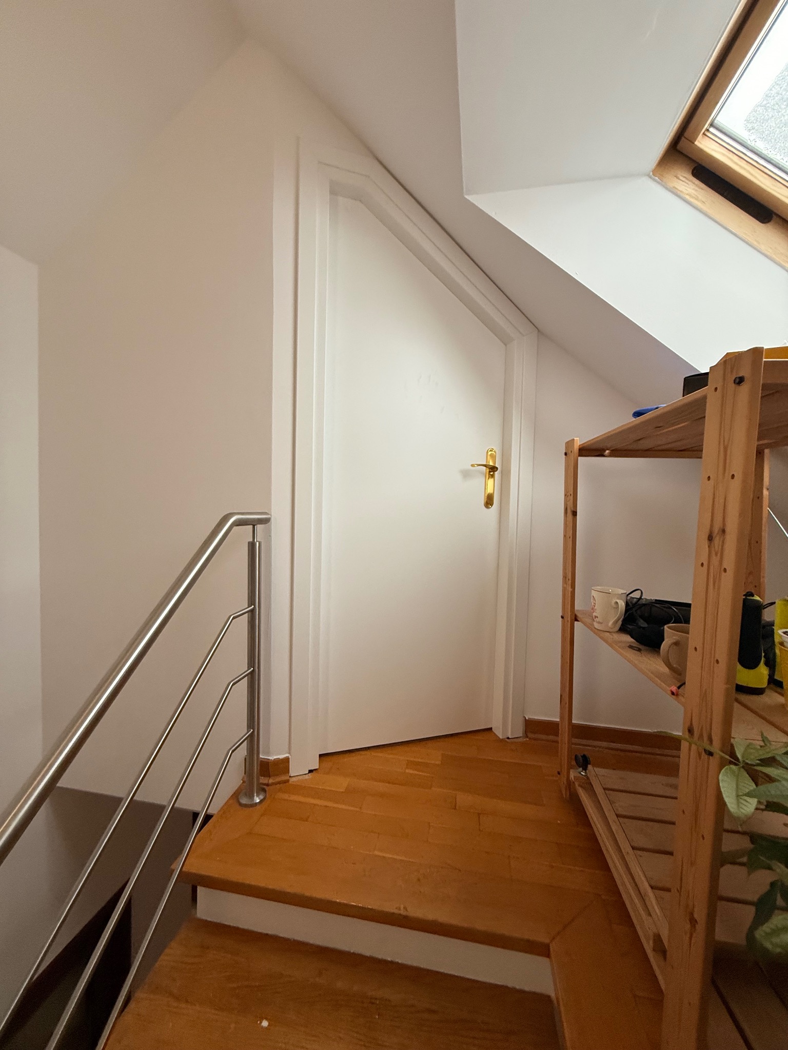 Białe drzwi z złotą klamką w skosie poddasza, widoczne schody z metalową barierką i drewniana podłoga. Ujęcie z dołu, widoczne półki z przedmiotami.