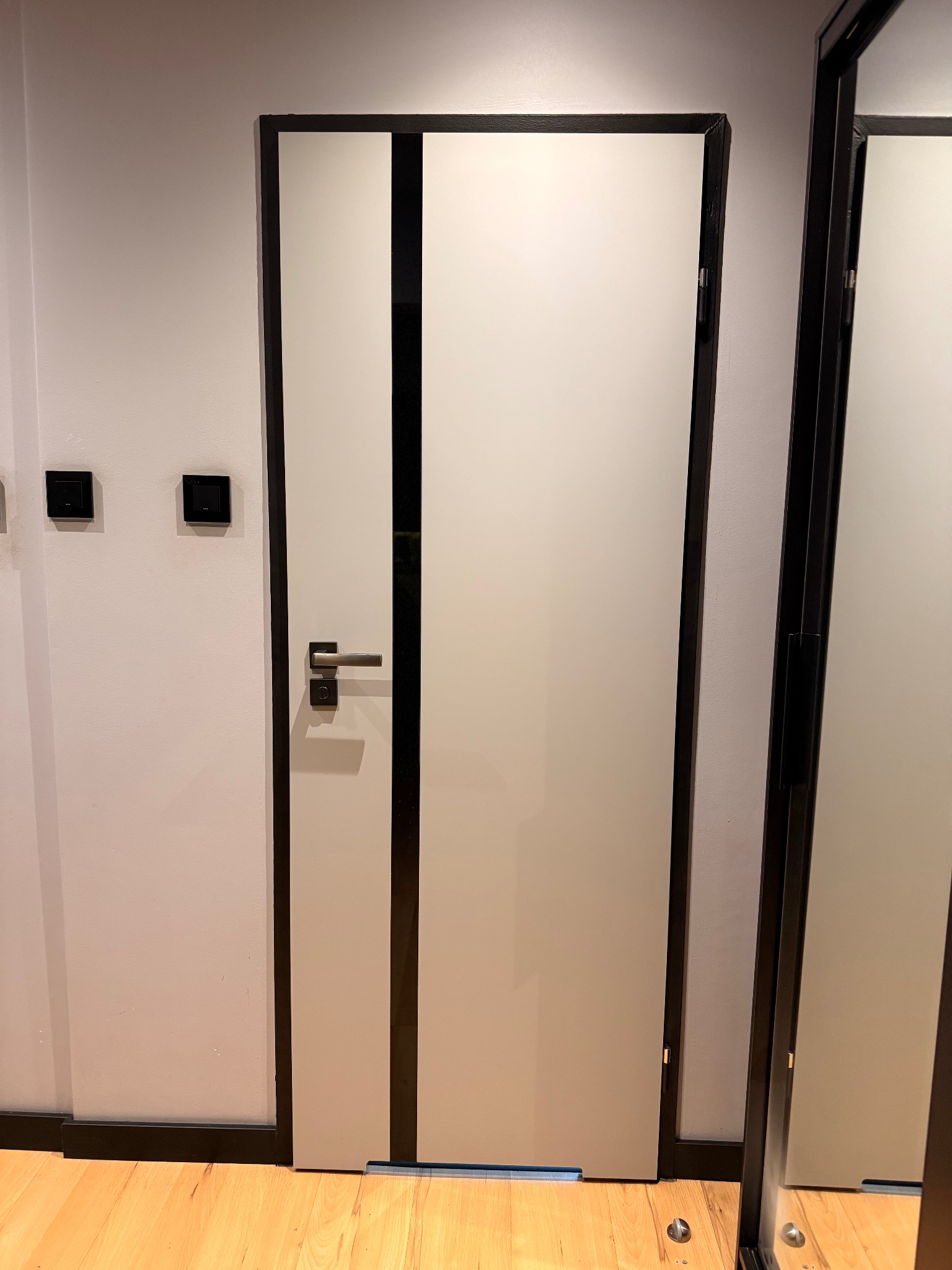 Zamknięte, nowoczesne drzwi wewnętrzne z czarnymi, pionowymi pasami na białym tle, widoczne na tle jasnej ściany i drewnianej podłogi. Minimalistyczny design.