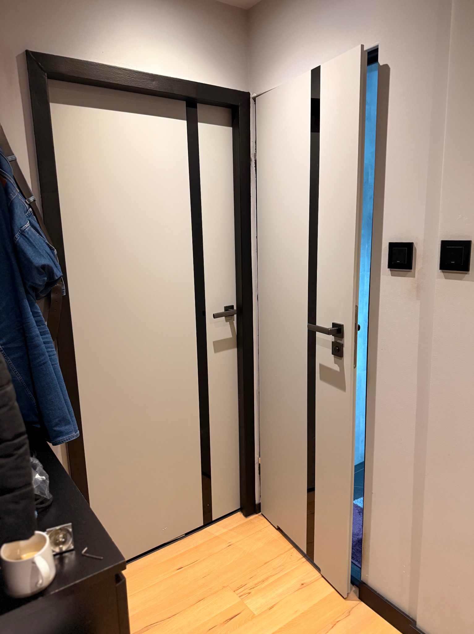 Nowoczesne, białe drzwi z czarnymi akcentami, zamontowane na starej futrynie. Widoczne dwie pary drzwi, jedna otwarta, ukazująca wnętrze. Na pierwszym planie fragment szafki z kubkiem.