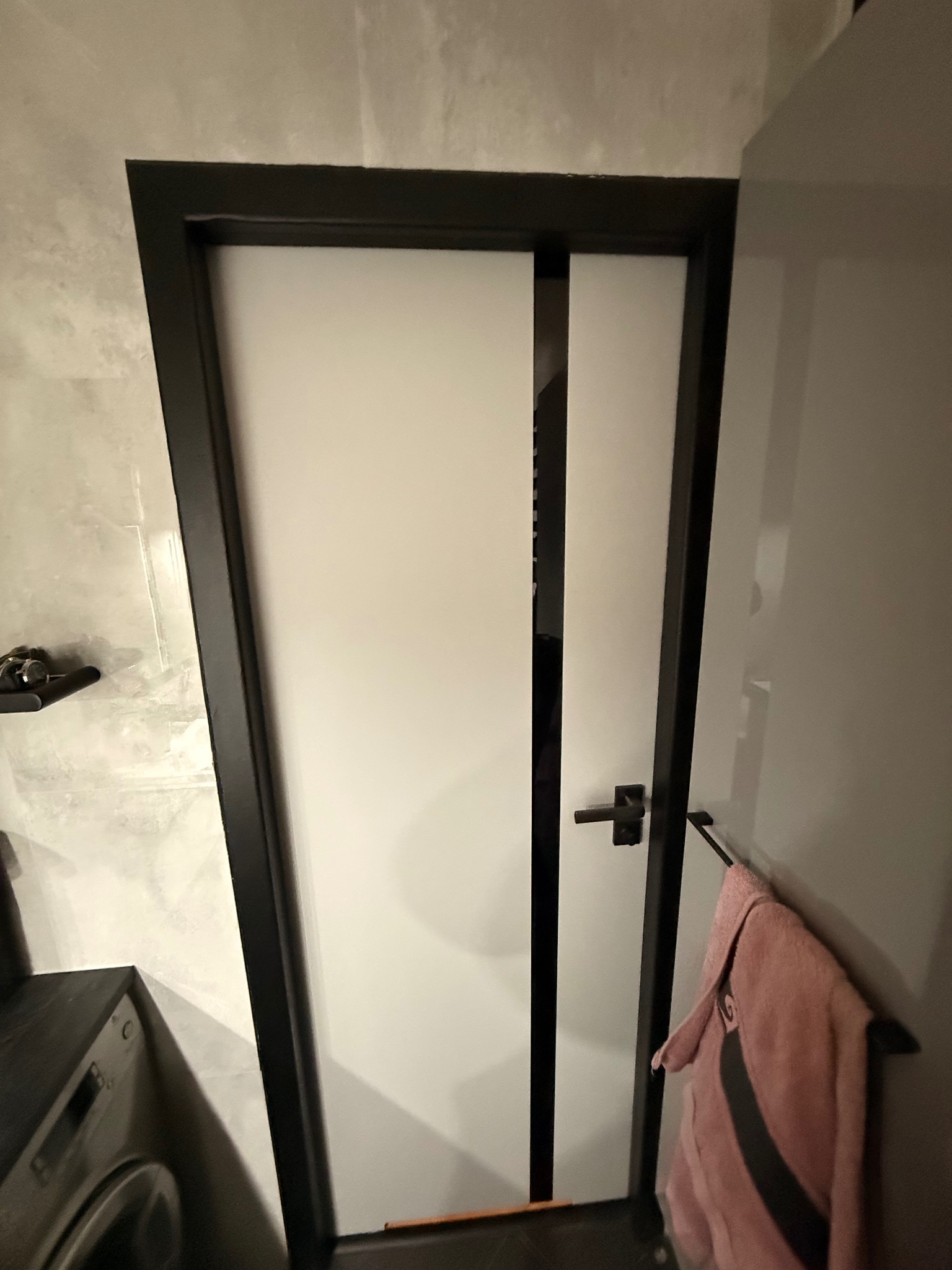 Nowoczesne, białe drzwi z czarną ramą i pionowym paskiem, zamontowane w łazience z szarą, marmurkową ścianą. Widoczny fragment pralki i ręcznik.