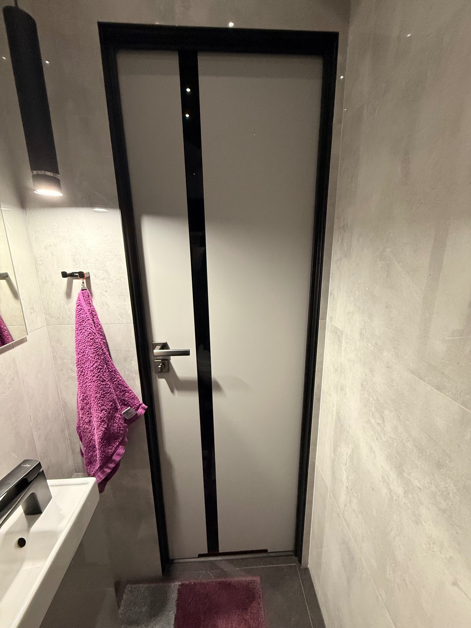 Nowoczesna łazienka z białą umywalką, fioletowym ręcznikiem i drzwiami z czarną ramą. Minimalistyczny design, jasne płytki, oświetlenie punktowe. Widok z dołu.