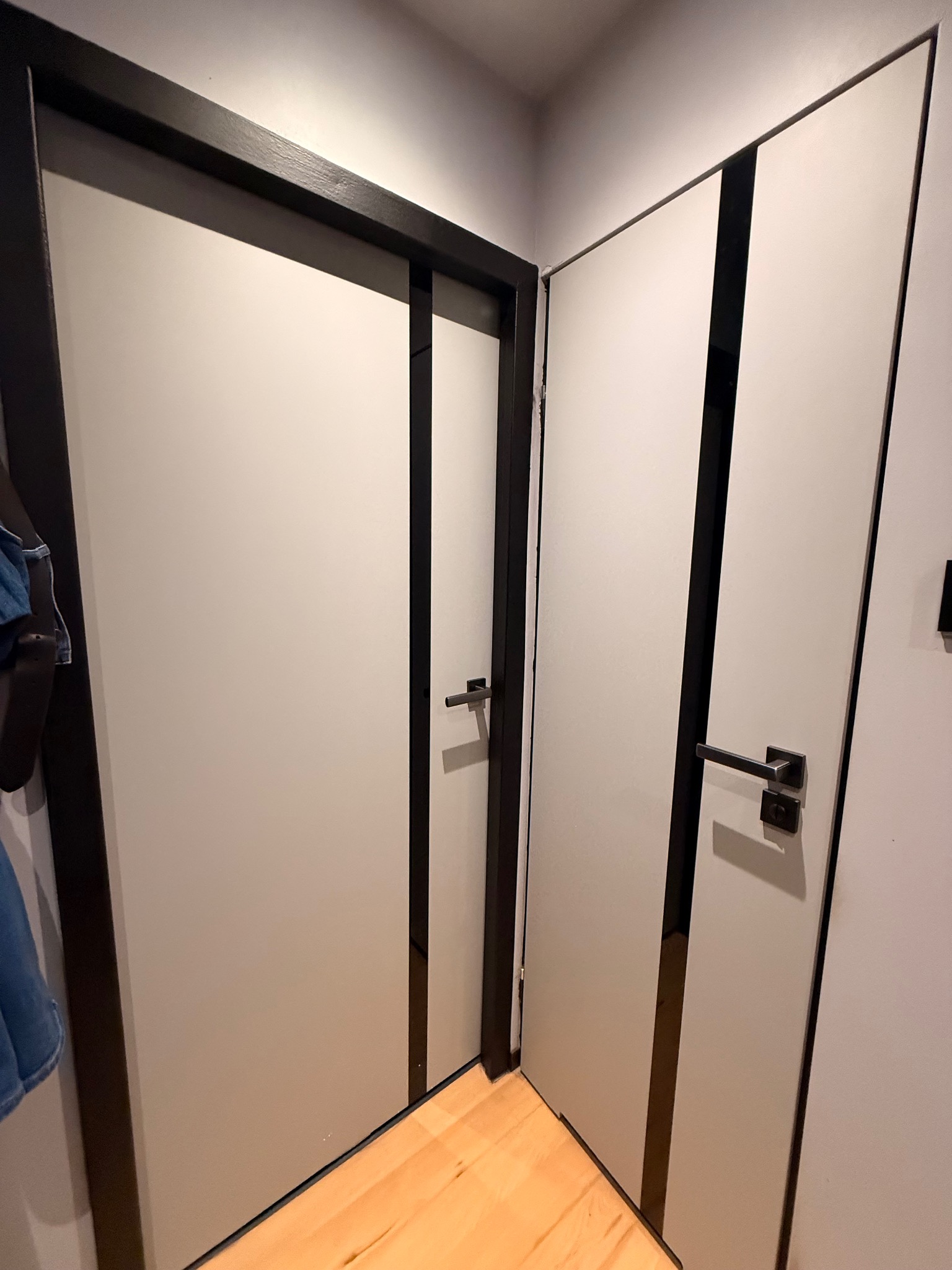 Dwie nowoczesne, szare drzwi z czarnymi ramami i klamkami na jasnej podłodze. Jedne drzwi są otwarte, drugie zamknięte. Minimalistyczny design.