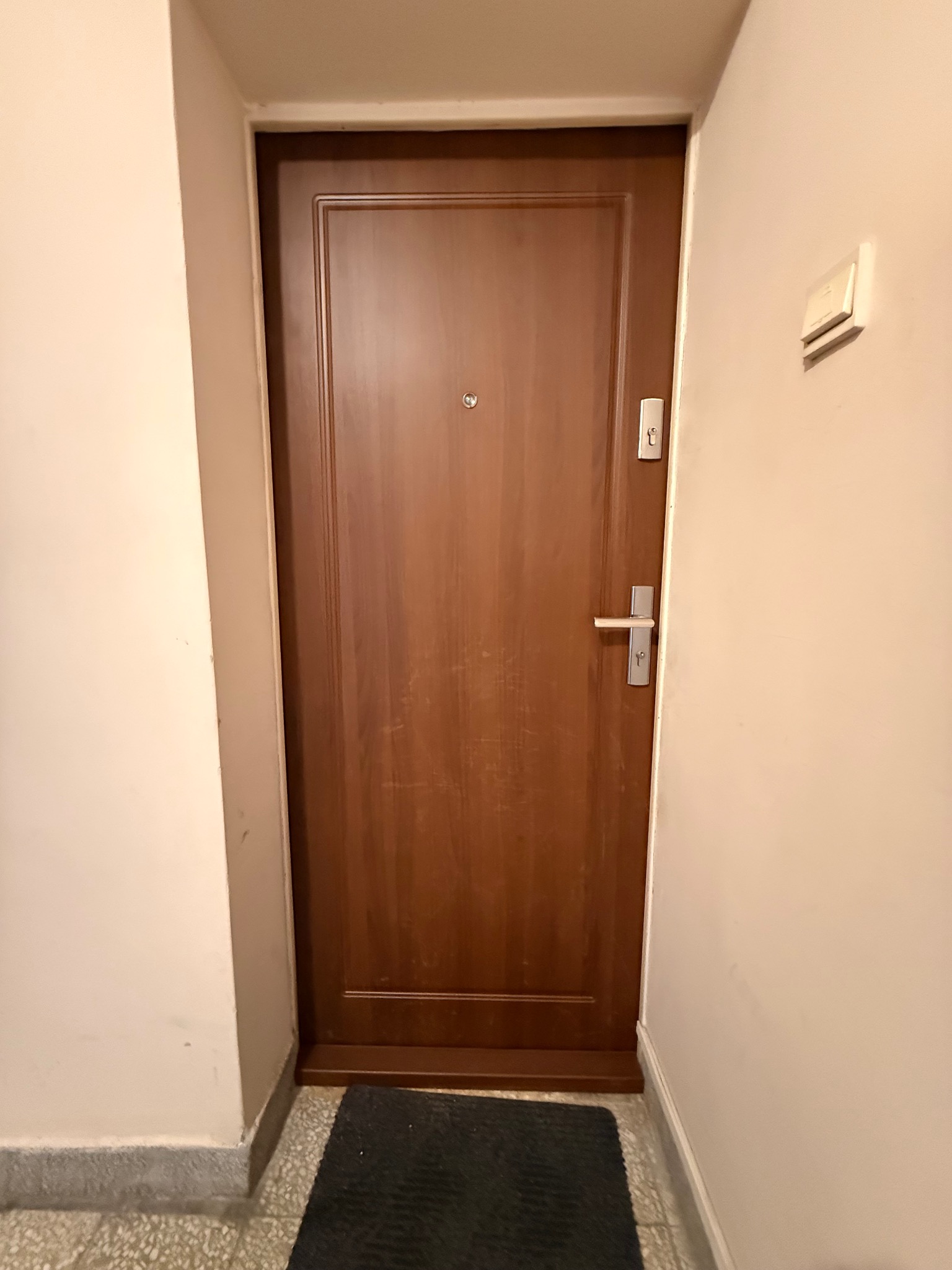 Zamknięte, brązowe drzwi wejściowe z wizjerem i srebrną klamką, na tle jasnej ściany z włącznikiem. Widoczny fragment wycieraczki i podłogi.