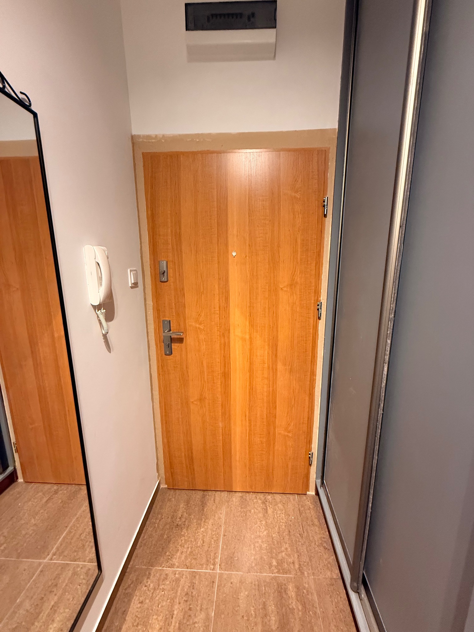 Jasne, drewniane drzwi wejściowe z metalową klamką i wizjerem, zamontowane w ościeżnicy. Obok ściana z domofonem i fragment lustra. Widoczna podłoga z płytek.