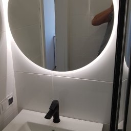 KINK-BUD - Nowoczesna łazienka: biała umywalka z czarną armaturą i okrągłym lustrem z podświetleniem LED, odbicie osoby w lustrze.