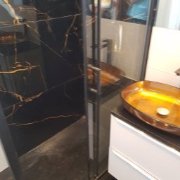 KINK-BUD - Nowoczesna łazienka z kabiną prysznicową z czarnymi płytkami imitującymi marmur ze złotymi żyłami i umywalką w kolorze bursztynowym, odbicie osób w lustrze.