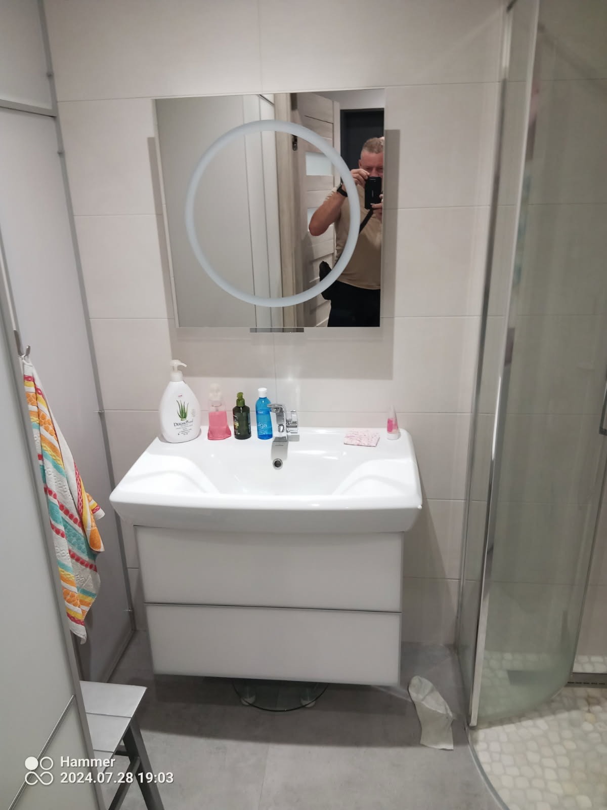 Biała umywalka z szafką w łazience, nad nią lustro z okrągłym podświetleniem, w którym odbija się osoba robiąca zdjęcie telefonem.