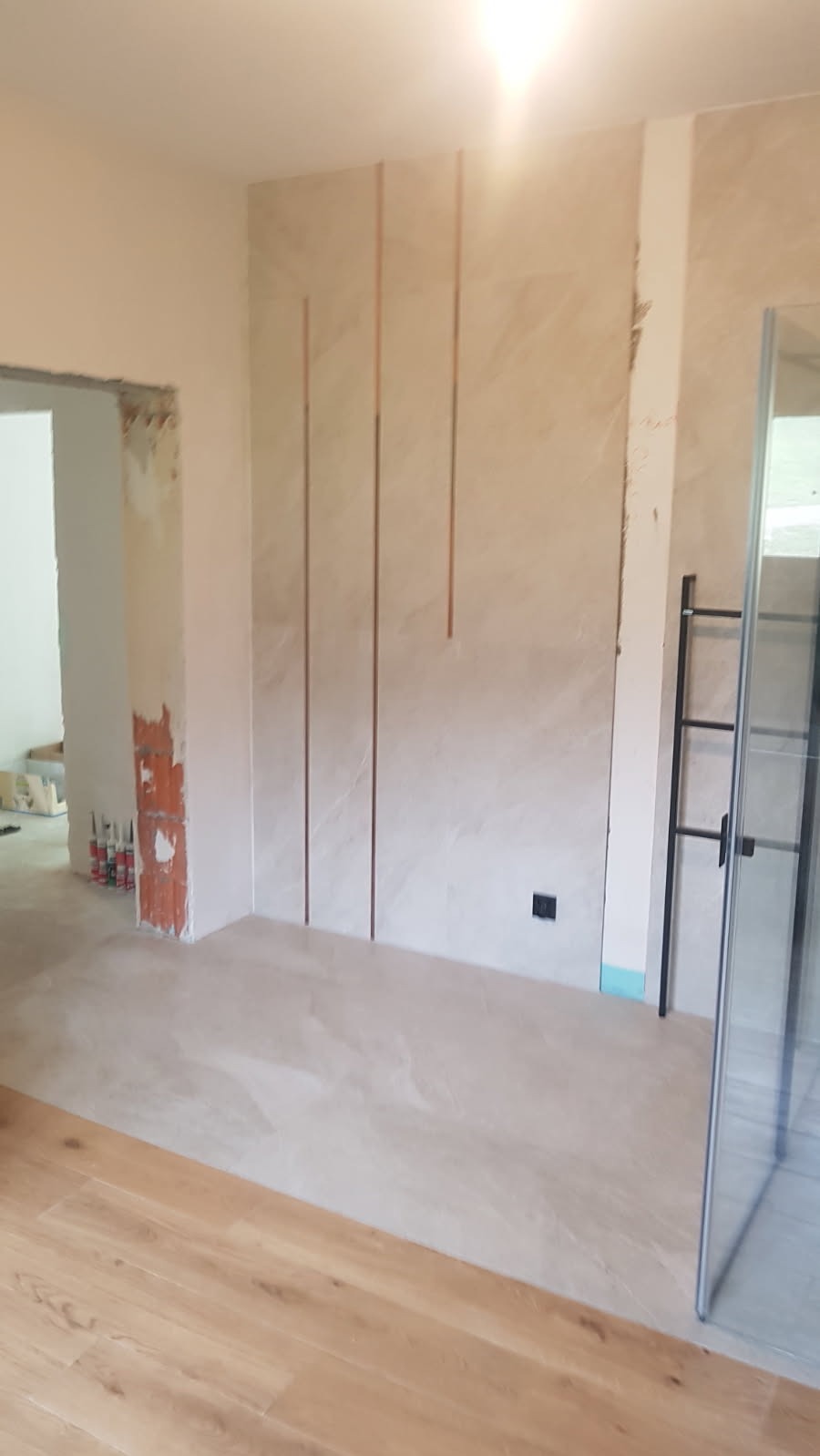 Niedokończona łazienka z płytkami imitującymi marmur na ścianie, pionowe metalowe listwy dekoracyjne, widoczne rury i kafelki pod prysznicem.