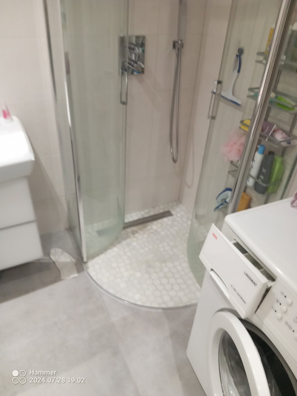 Nowoczesna kabina prysznicowa z zaokrąglonym szkłem, białe heksagonalne płytki na podłodze, pralka AEG Lavamat w rogu łazienki.