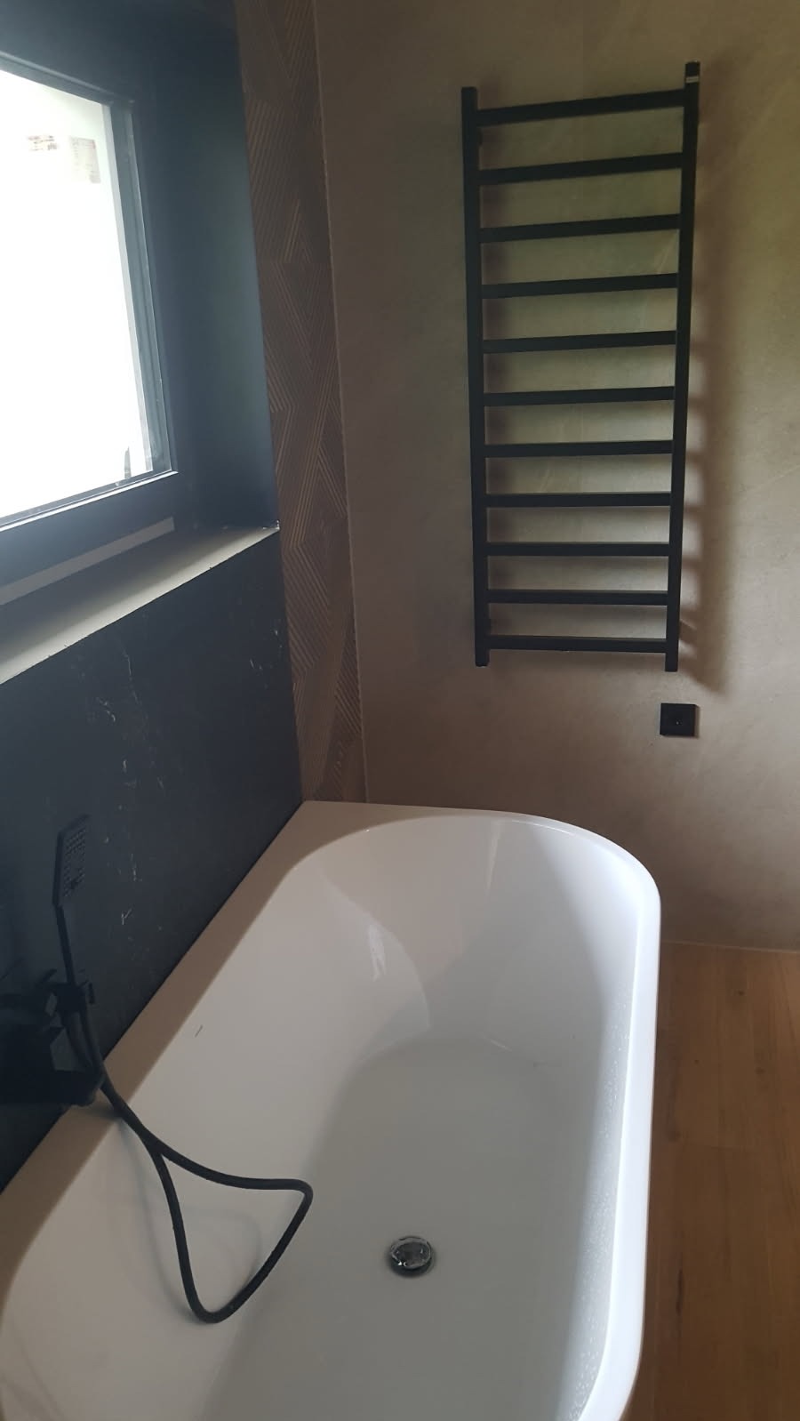 Biała wanna wolnostojąca z czarną armaturą i grzejnikiem drabinkowym w łazience z akcentami kamienia i drewna.