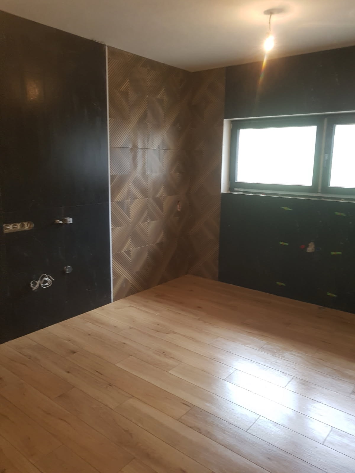 Wnętrze łazienki w trakcie remontu z położonymi panelami podłogowymi, częściowo wykończonymi ścianami z czarnych i wzorzystych brązowych płytek, widoczne puszki elektryczne.