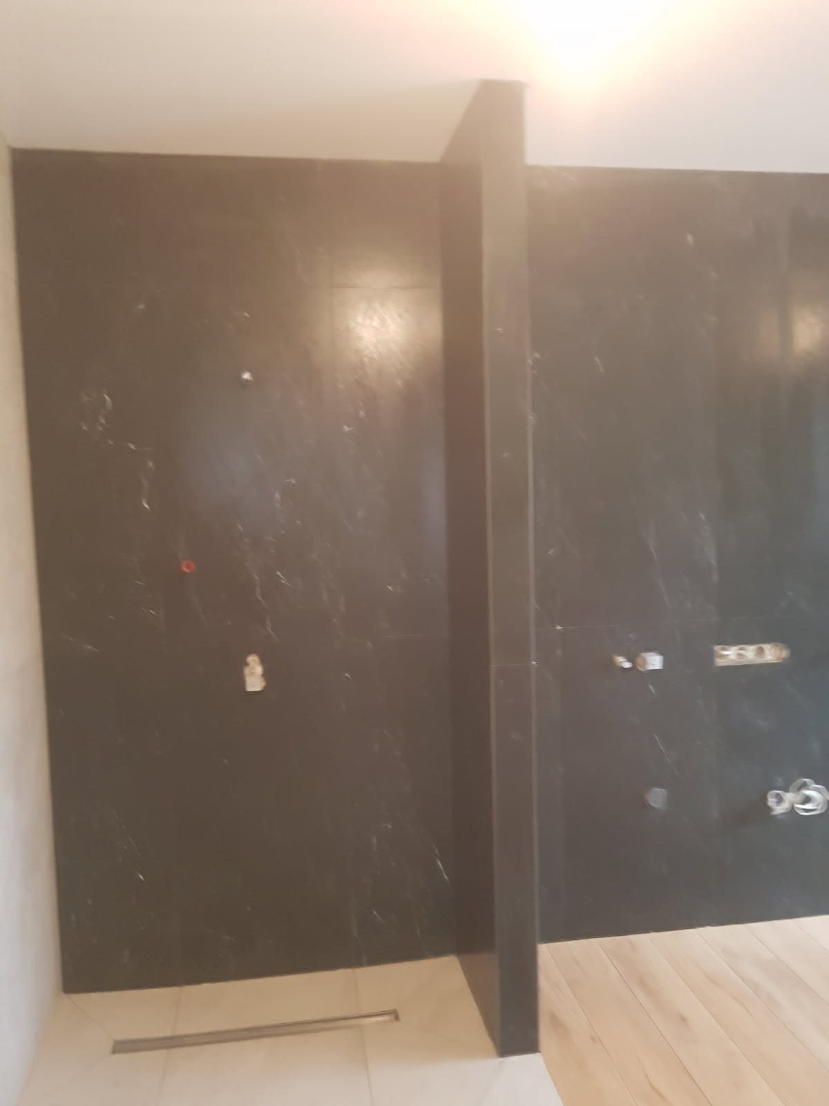 Wykończona kabina prysznicowa z ciemnymi, marmurkowymi płytkami, widoczne odpływy liniowe i przygotowane punkty instalacyjne na ścianie.