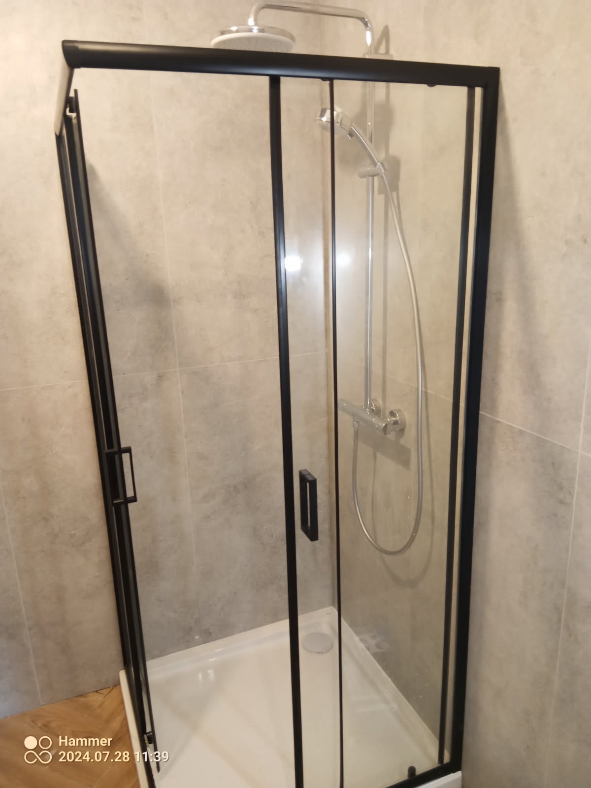 Nowoczesna kabina prysznicowa z czarną ramą, widok z boku, szklane drzwi, biały brodzik, chromowana bateria i deszczownica.