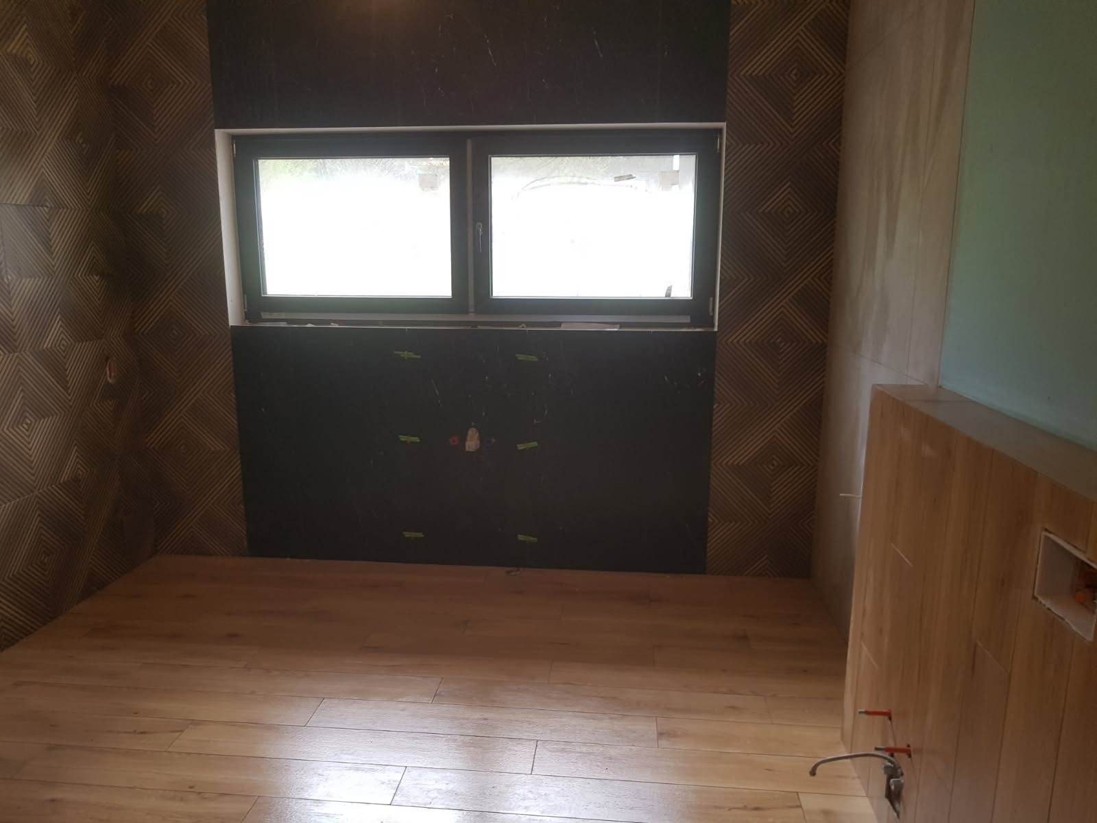 Łazienka w trakcie wykańczania: ułożona podłoga z paneli imitujących drewno, fragment ściany z płytek o geometrycznym wzorze, czarna ściana z widocznymi otworami na instalacje oraz okno.