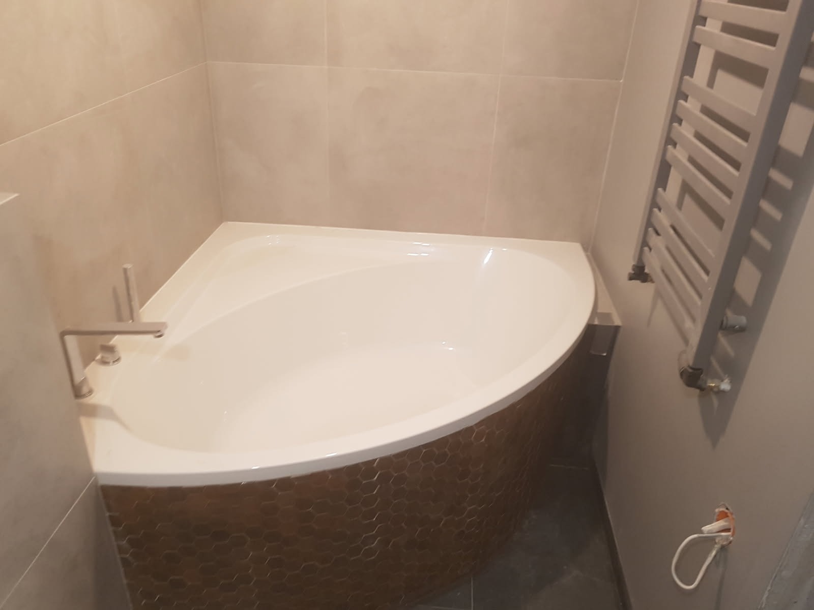 Narożna wanna z obudową z mozaiki w odcieniach brązu, chromowana bateria, grzejnik drabinkowy na ścianie w łazience wykończonej beżowymi płytkami.