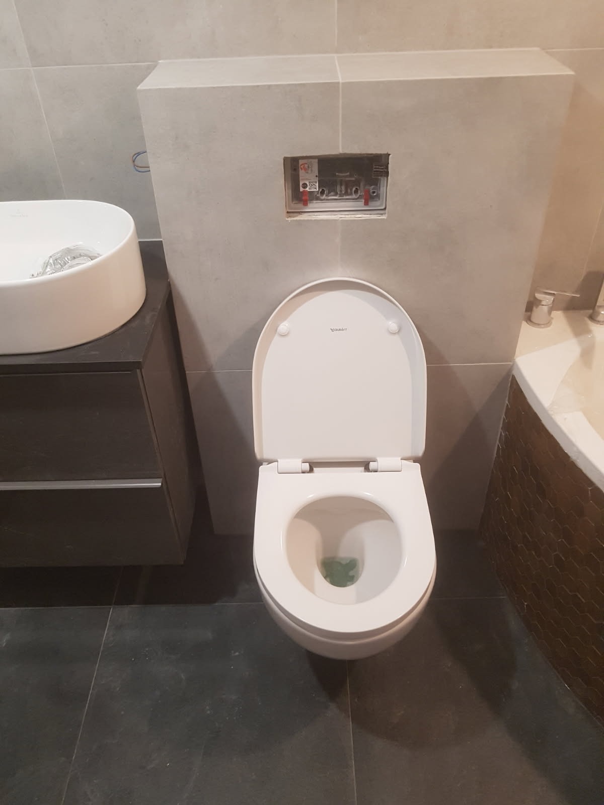 Nowoczesna toaleta Duravit z otwartą deską, zamontowana na tle szarych płytek, z widoczną instalacją podtynkową i umywalką nablatową po lewej stronie.