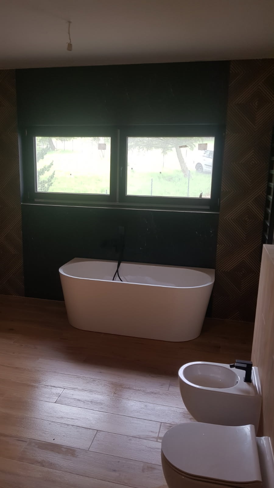 Nowoczesna łazienka z wolnostojącą wanną, bidetem i toaletą w jasnych barwach, wykończona płytkami imitującymi drewno i czarnym kamieniem, z widokiem na zewnątrz przez okno.