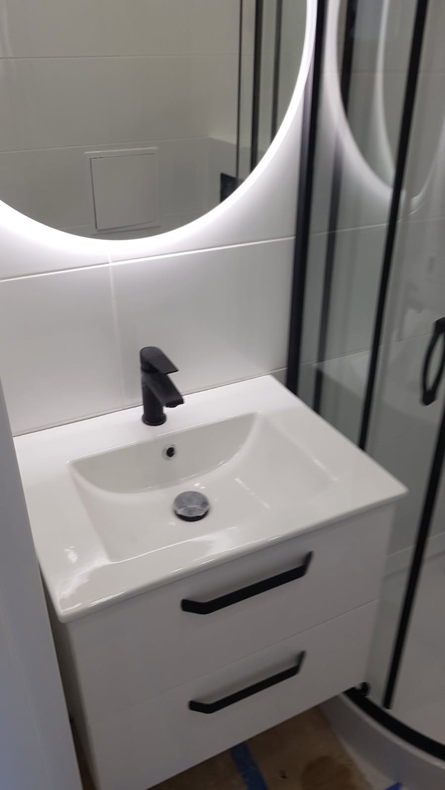 Biała umywalka z czarną baterią i szafką z dwoma szufladami w nowoczesnej łazience z okrągłym lustrem podświetlanym LED i kabiną prysznicową z czarnymi profilami.
