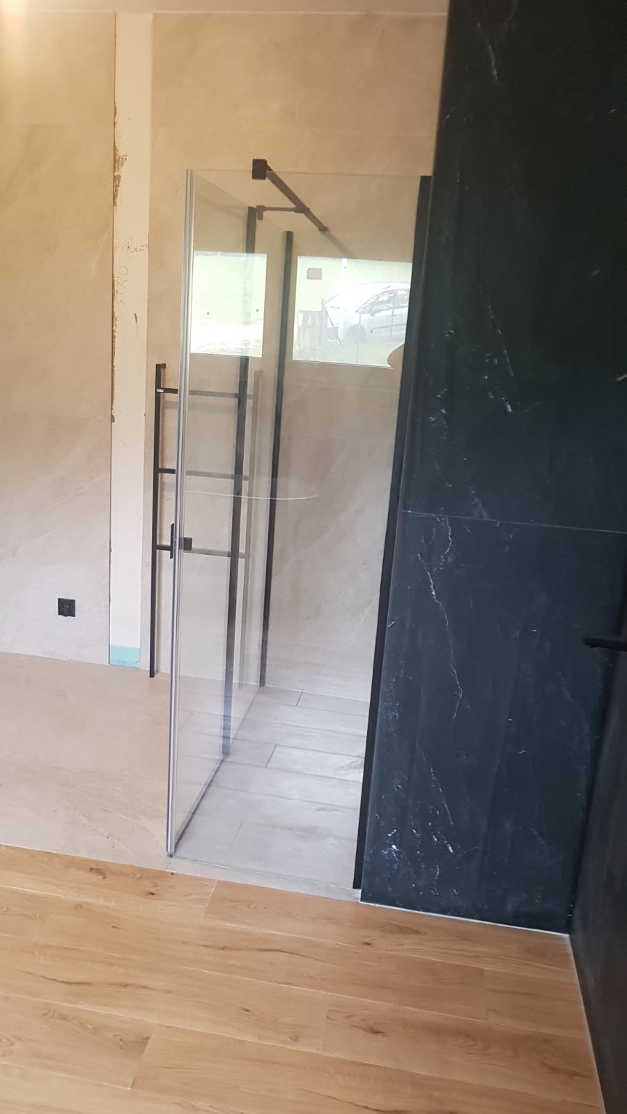 Szklana kabina prysznicowa z czarnymi profilami w trakcie montażu, widoczne niedokończone ściany i podłoga, jasne płytki imitujące marmur i ciemne płytki imitujące kamień.