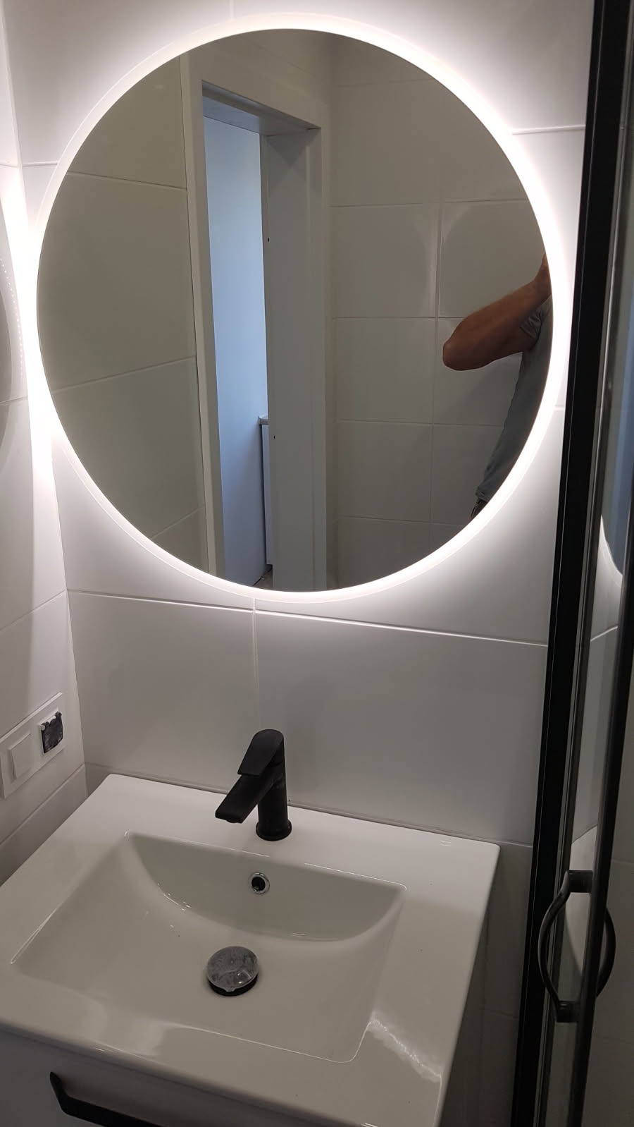 Nowoczesna łazienka: biała umywalka z czarną armaturą i okrągłym lustrem z podświetleniem LED, odbicie osoby w lustrze.