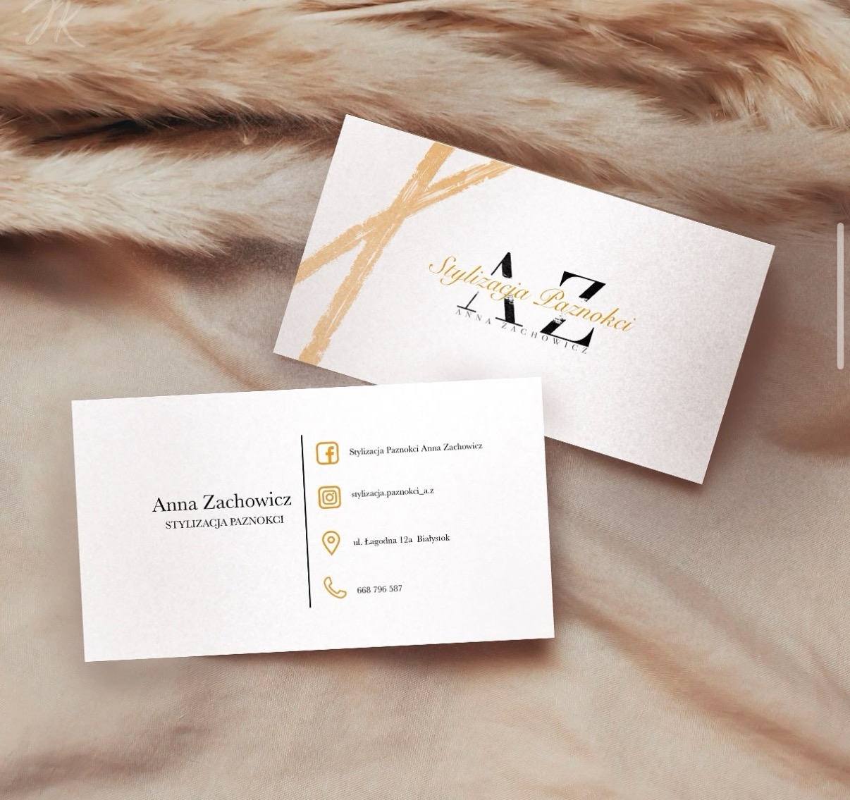 Dwie wizytówki firmy stylizującej paznokcie, z minimalistycznym designem w odcieniach bieli i złota, ułożone na tle z beżowych piór.