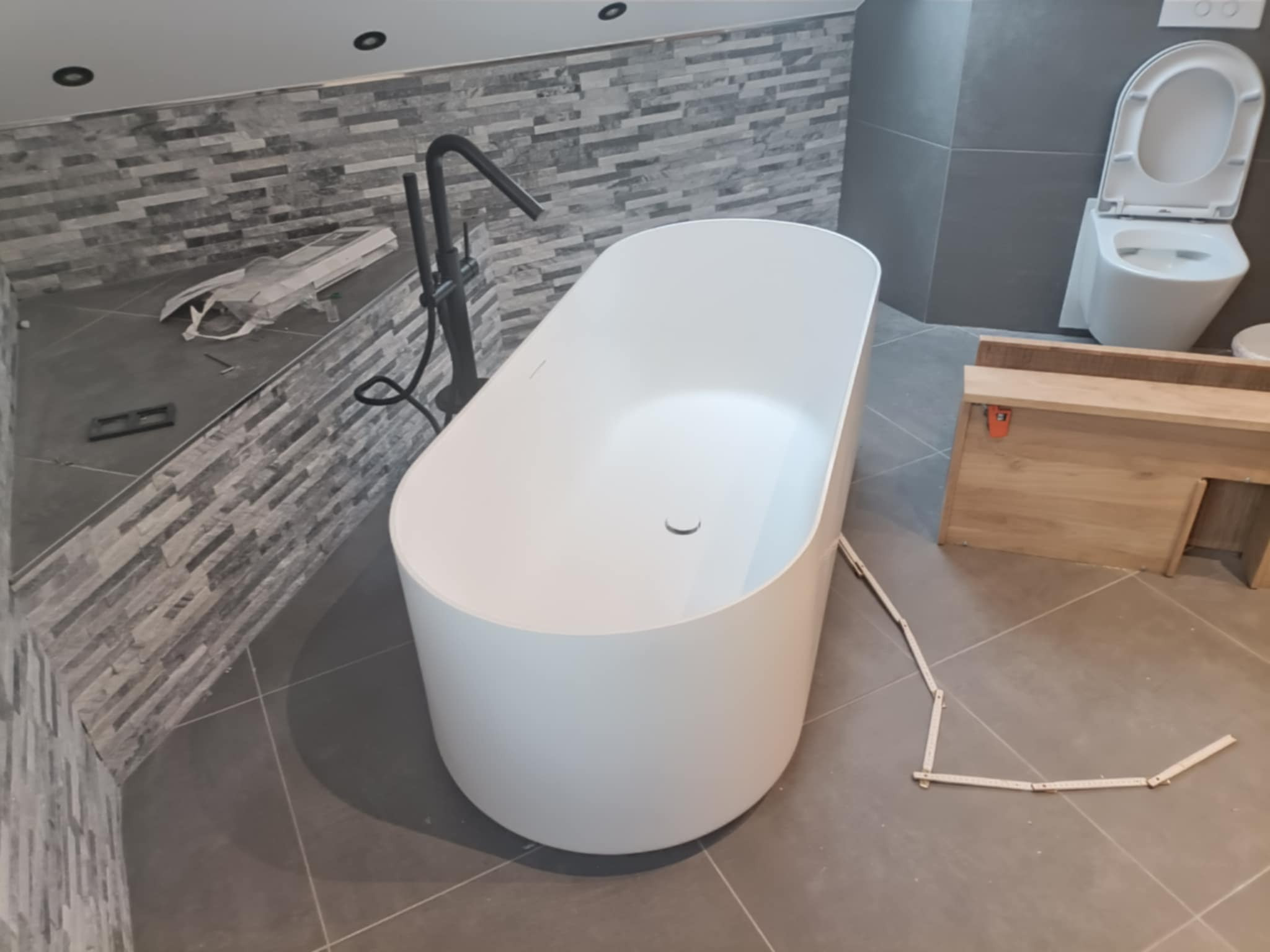 Nowoczesna, wolnostojąca wanna owalna z czarną armaturą, szara ściana z płytek imitujących kamień, częściowo ułożone płytki pod prysznicem, otwarta toaleta podwieszana i drewniana szafka w trakcie...