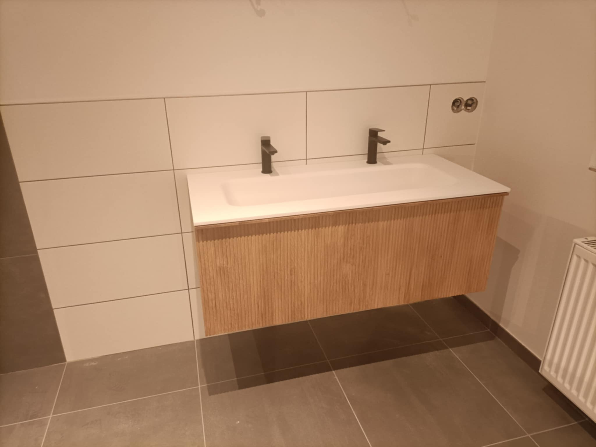 Nowoczesna łazienka z podwójną umywalką nablatową i drewnianą szafką wiszącą, czarne baterie, białe płytki prostokątne na ścianie i szara podłoga