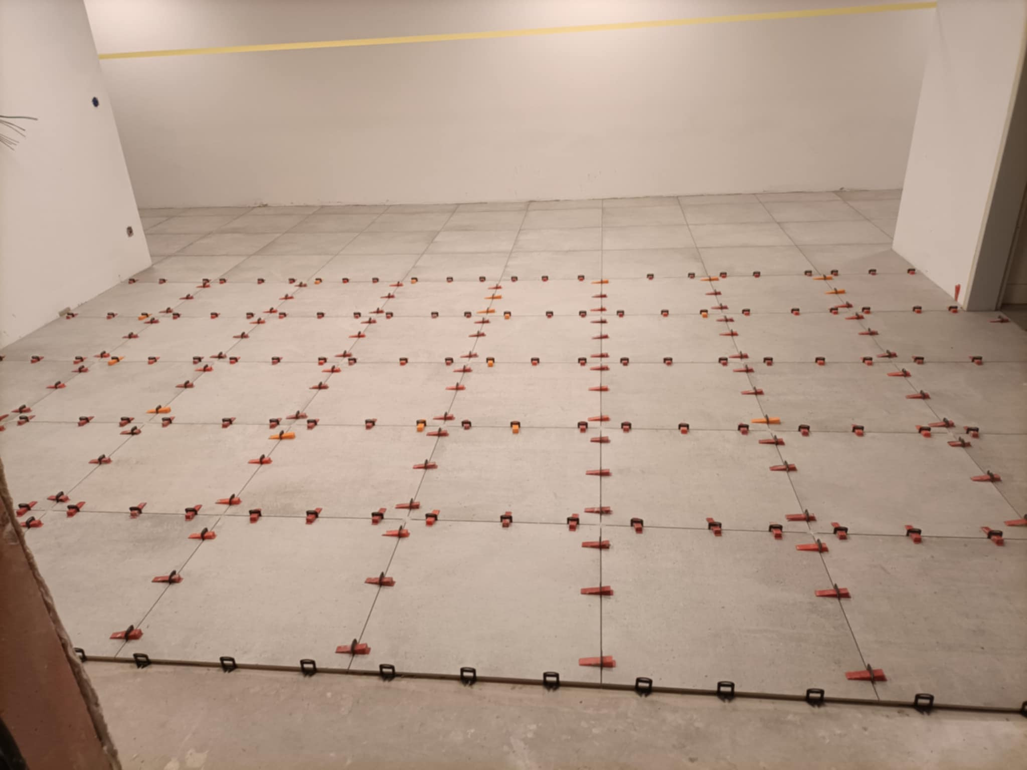 Układanie jasnych płytek podłogowych z użyciem systemu poziomowania płytek, widoczne klipsy i kliny w kolorze pomarańczowym i czerwonym, taśma malarska na ścianie.