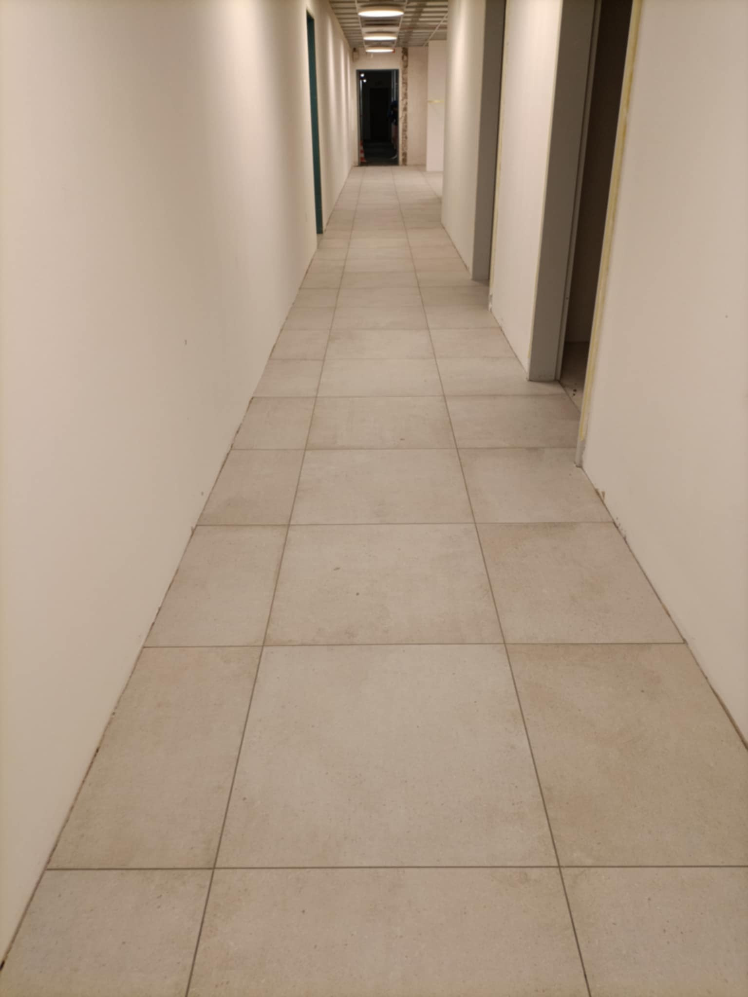 Długi korytarz wyłożony kwadratowymi, beżowymi płytkami podłogowymi, z białymi ścianami i widocznymi otworami drzwiowymi po obu stronach.