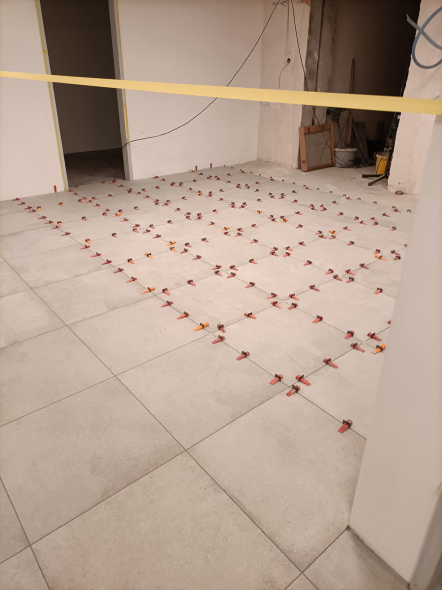 Jasnoszare płytki podłogowe w trakcie układania, z systemem poziomowania z czerwonymi i pomarańczowymi klinami, widoczne kable i żółta taśma zabezpieczająca w tle.
