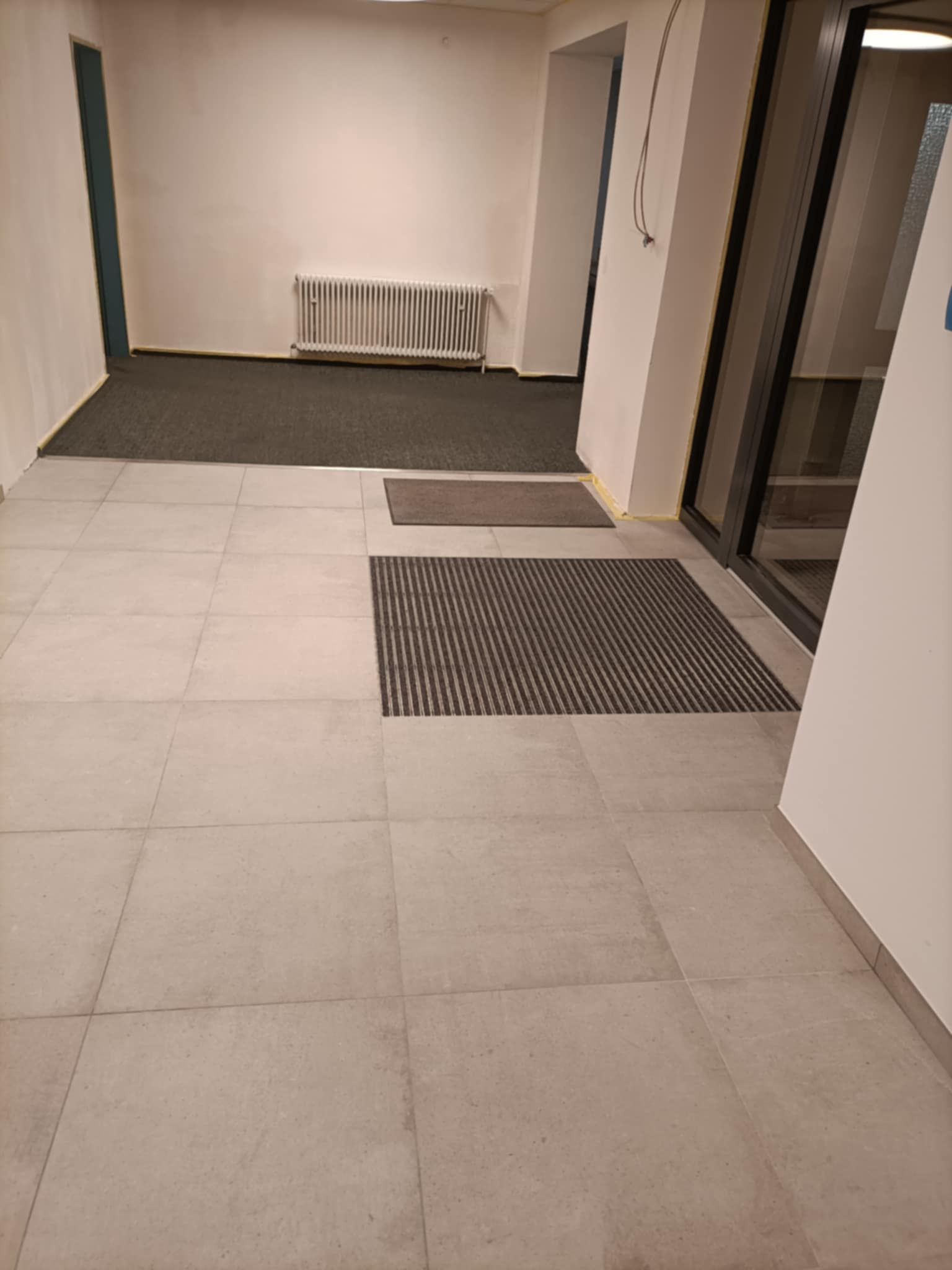 Korytarz wyłożony jasnoszarymi, kwadratowymi płytkami podłogowymi, z widocznymi wycieraczkami przy wejściu i przejściu do pomieszczenia z wykładziną oraz grzejnikiem.