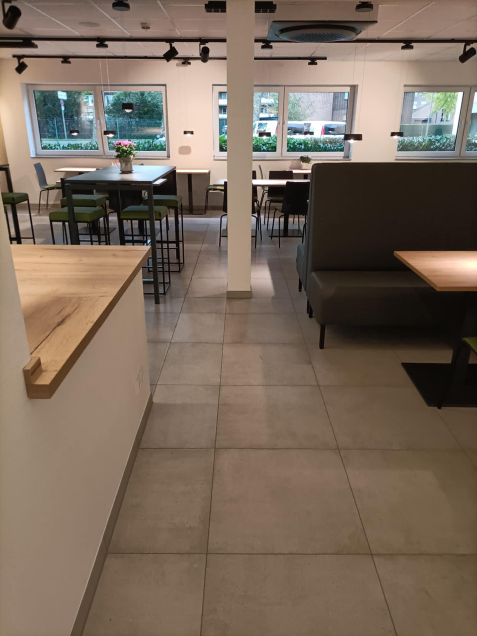 Wnętrze biurowe z szarymi płytkami podłogowymi, stołami z zielonymi krzesłami, szarą sofą i drewnianym blatem, oświetlone reflektorami sufitowymi.