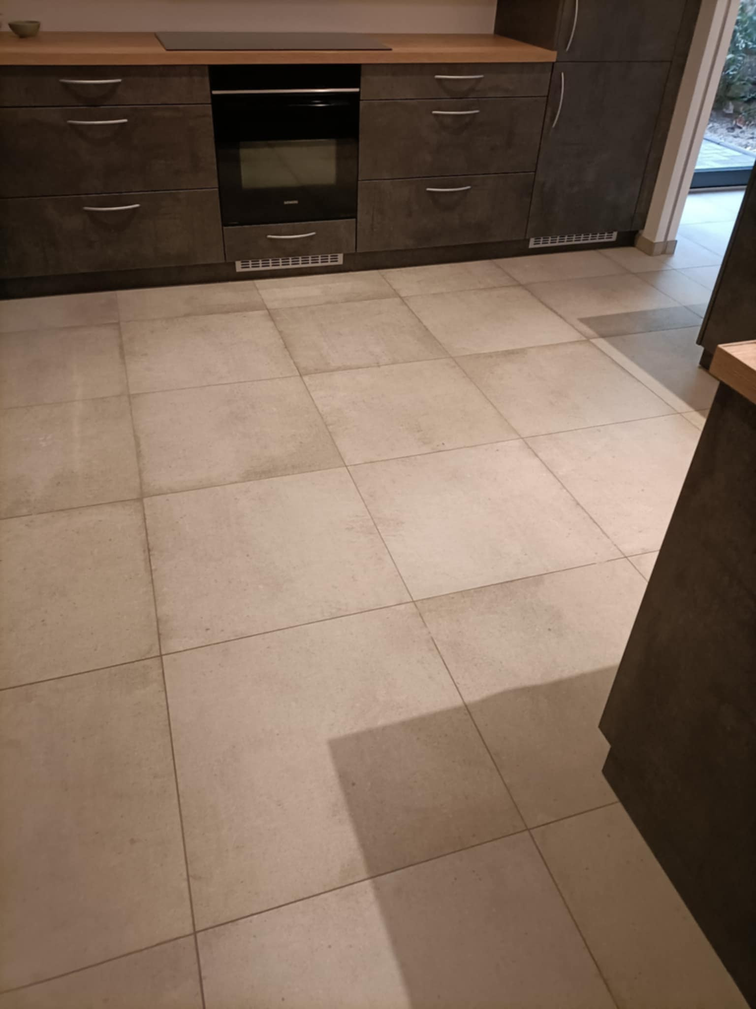 Beżowe, kwadratowe płytki podłogowe w kuchni, ułożone symetrycznie, widoczne szafki kuchenne z ciemnego drewna w tle.