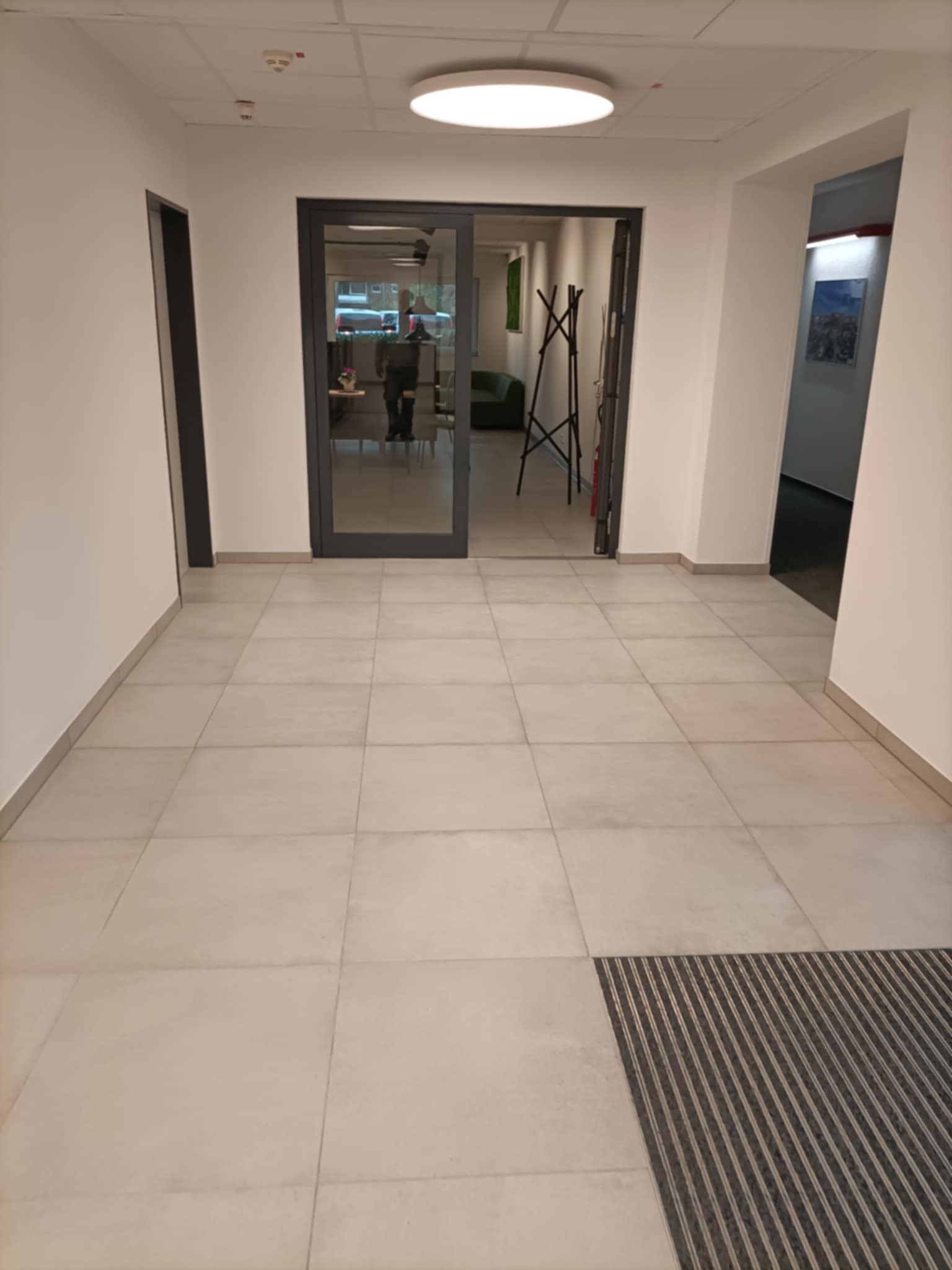 Jasne, kwadratowe płytki podłogowe w korytarzu biurowym, widok na szklane drzwi z ciemnymi ramami i wycieraczkę przy wejściu.