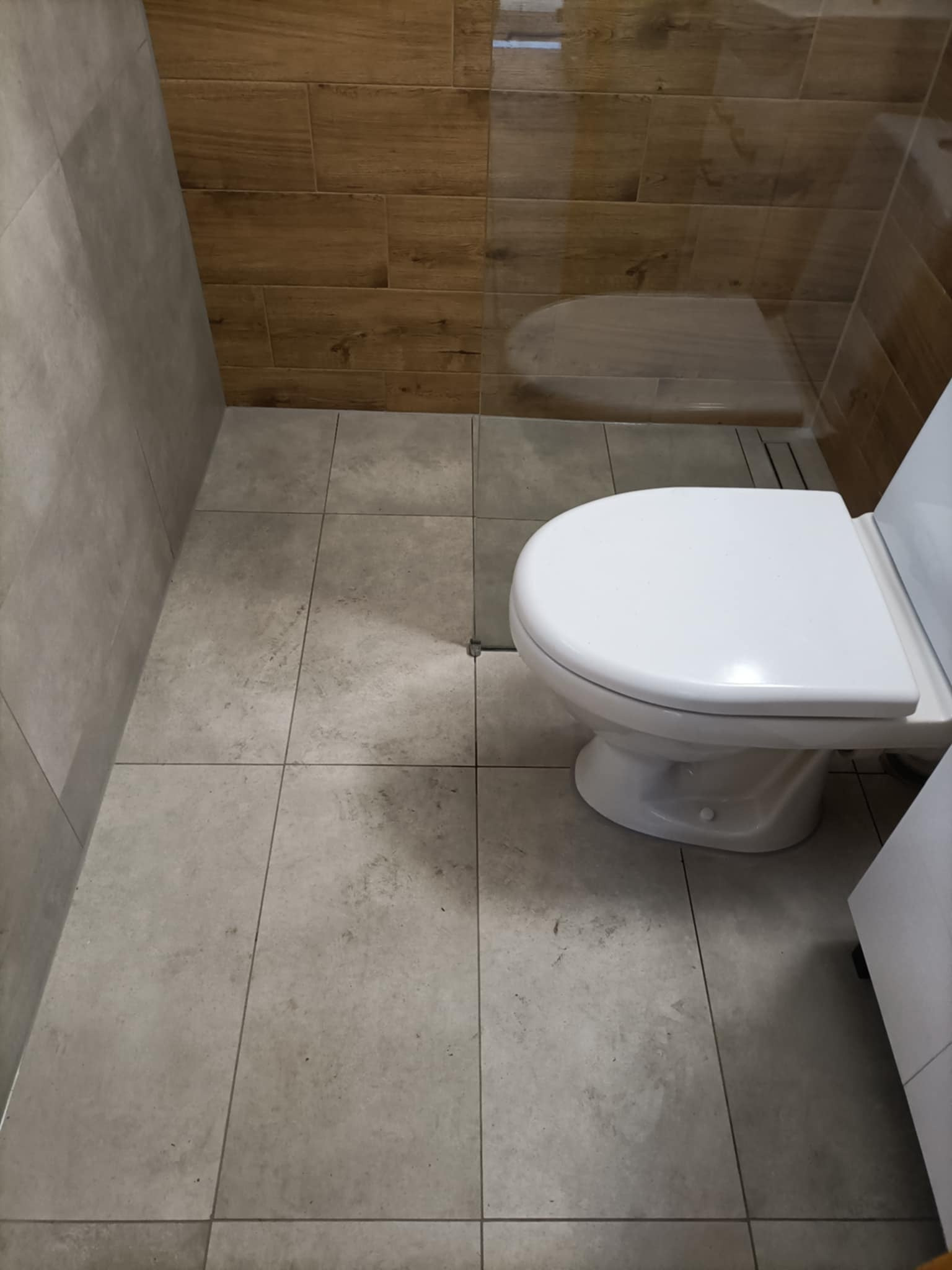 Wykończona łazienka z toaletą, szarą podłogą i drewnopodobnymi płytkami na ścianie. Widoczny odpływ liniowy.