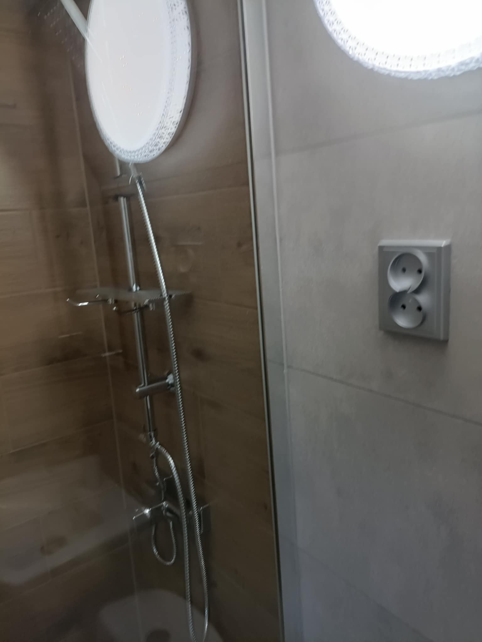 Nowoczesna kabina prysznicowa z deszczownicą i półkami, wykończona płytkami imitującymi drewno i szarymi płytkami. Widoczne gniazdko elektryczne.