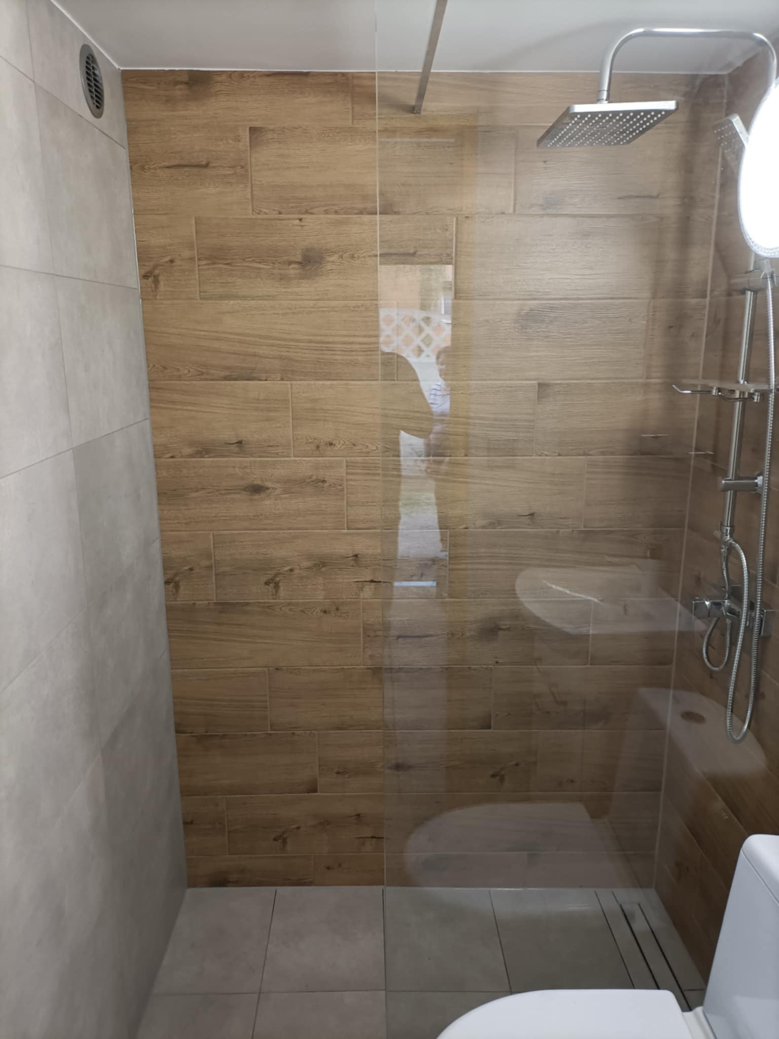 Nowoczesna kabina prysznicowa z drewnopodobnymi płytkami i szklaną ścianką. Widoczna armatura i odpływ liniowy. Minimalistyczny design, jasne płytki na podłodze.